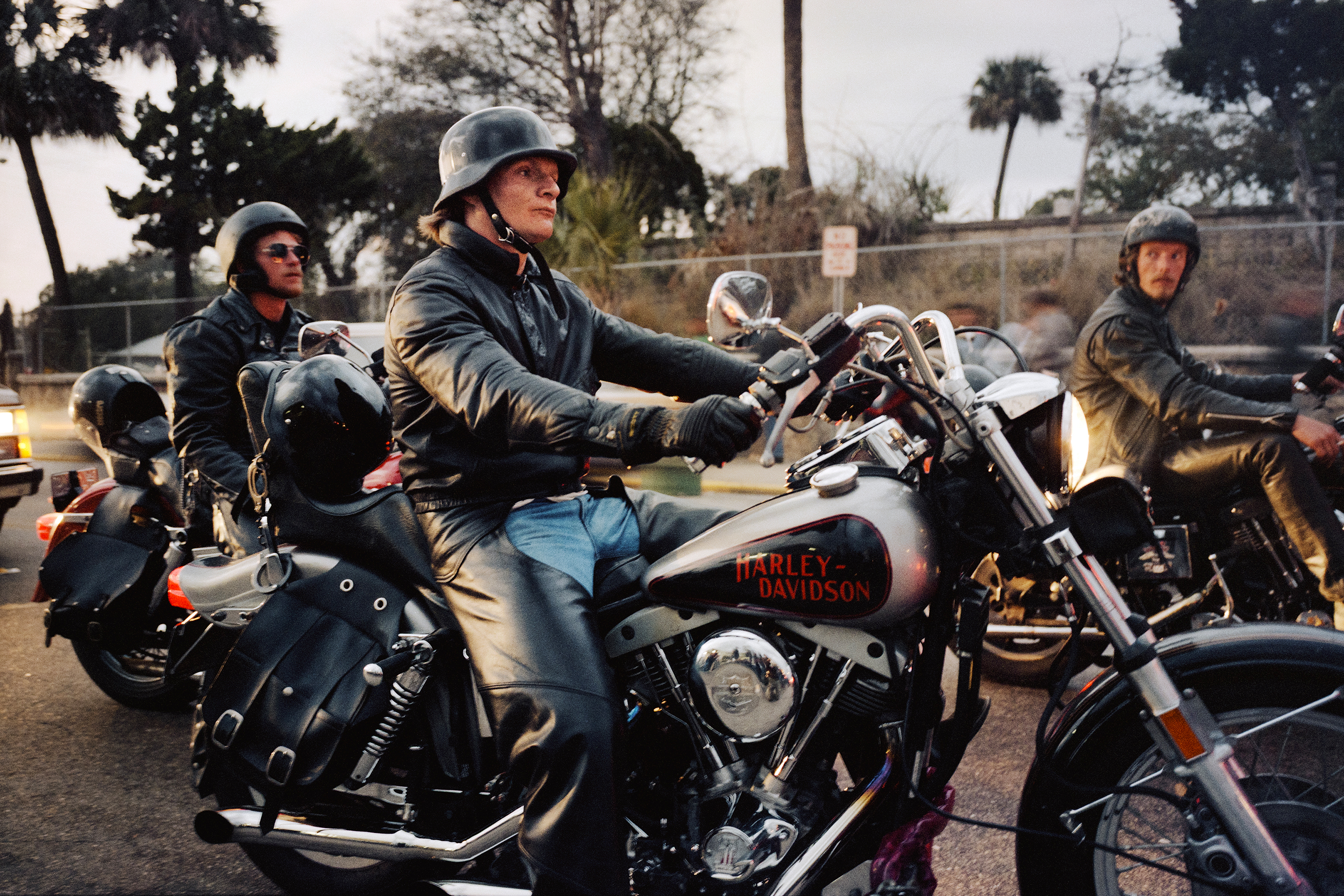 Three motorcyclists in leather jackets and helmets sit on Harley-Davidson bikes in sunny outdoor setting with palm trees visible.