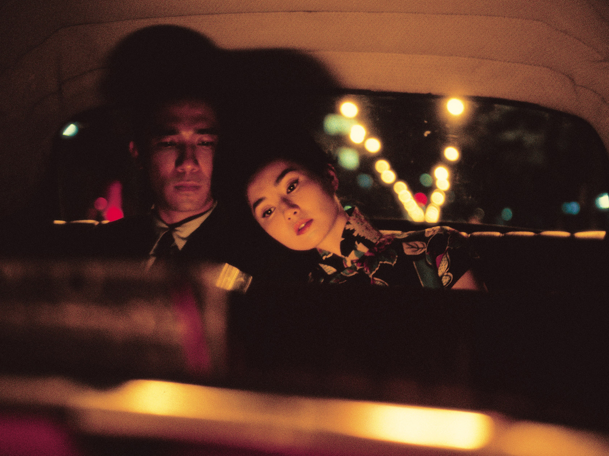 Tony Leung and Maggie Cheung sitting in the backseat of a car at night, illuminated by the glow of lights in the background.