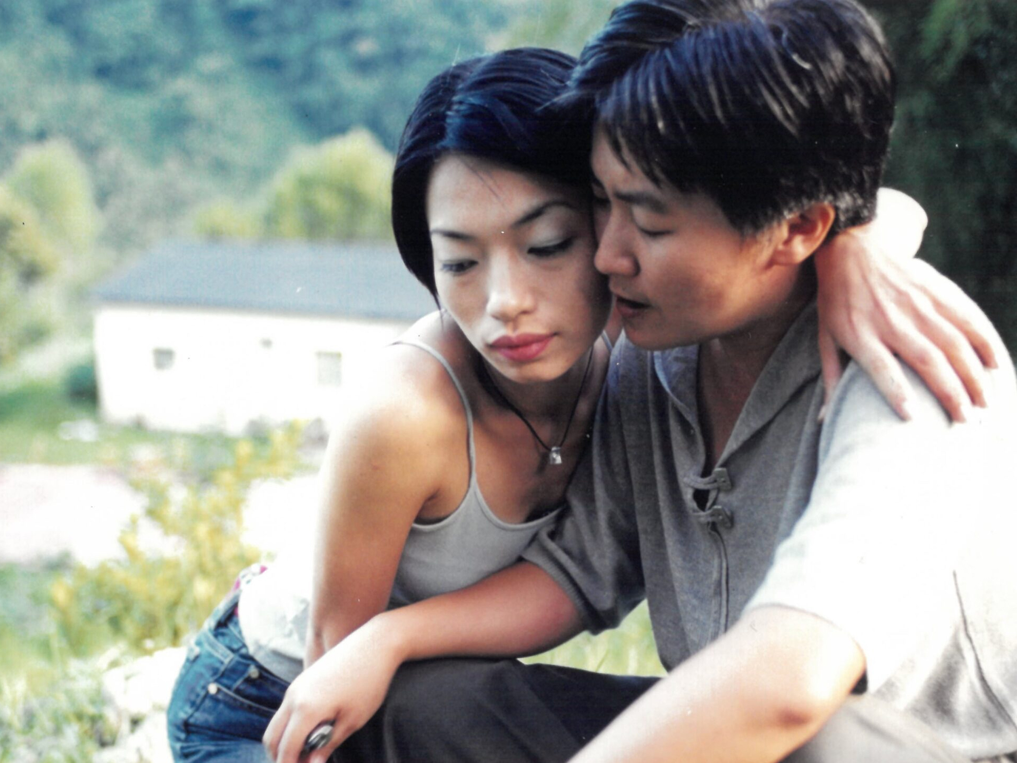 Two young East Asian adults, a man and a woman, sit close together in a grassy field. The woman has her arm around the man's shoulder.