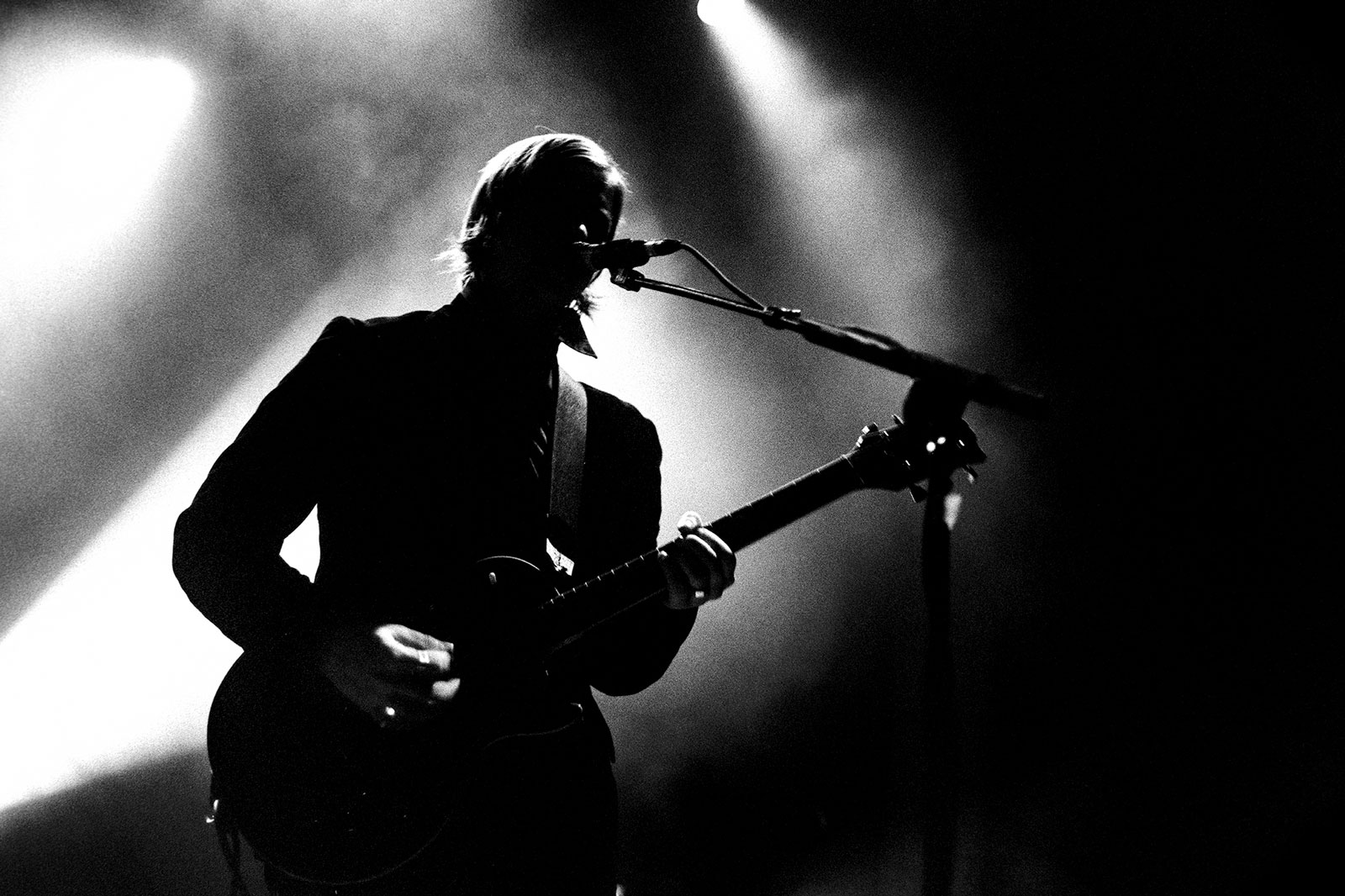 Black and white silhouette of guitarist with microphone against bright stage lights and smoke.