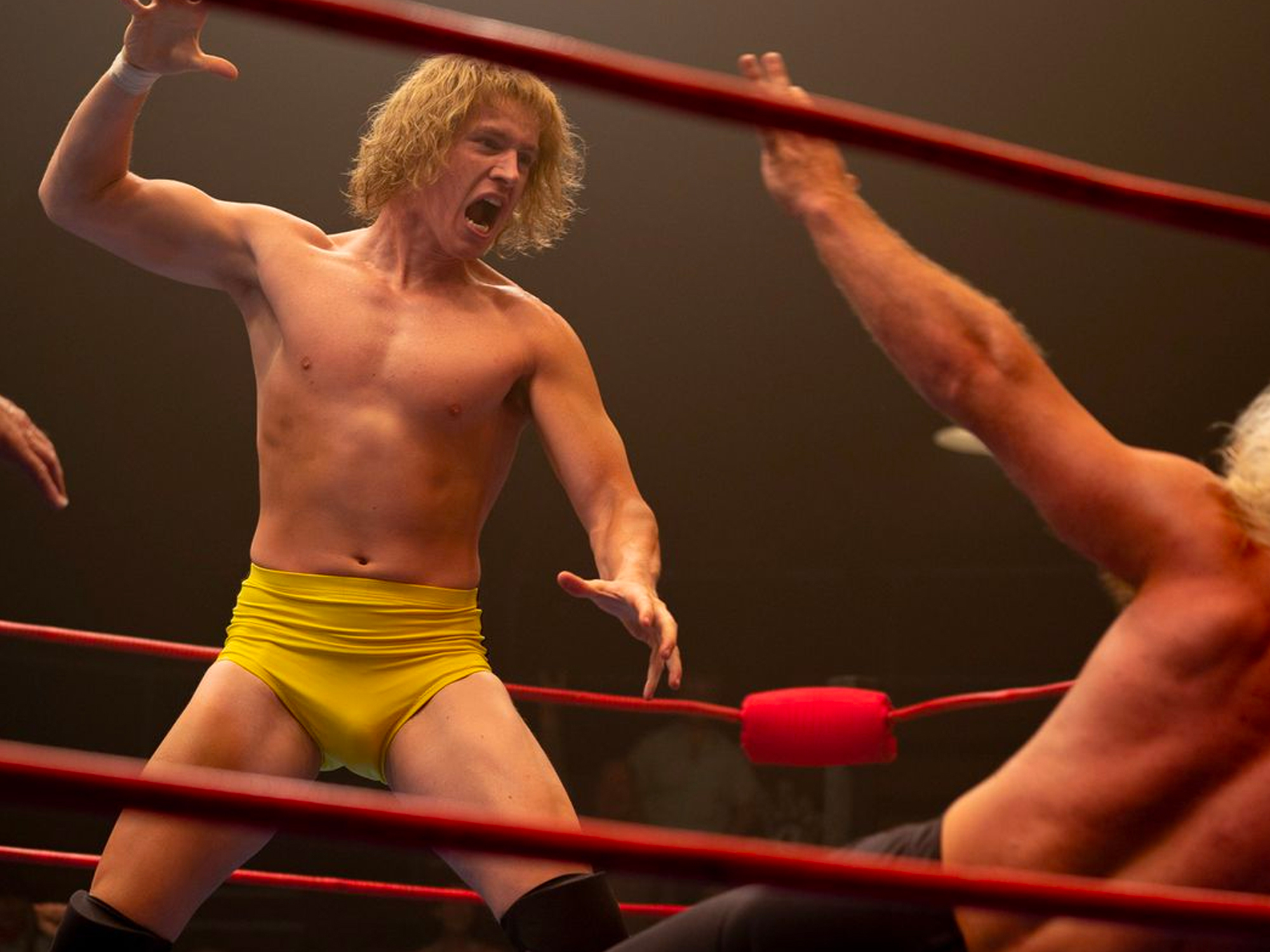 Muscular male wrestler in yellow trunks raising arms in wrestling ring.