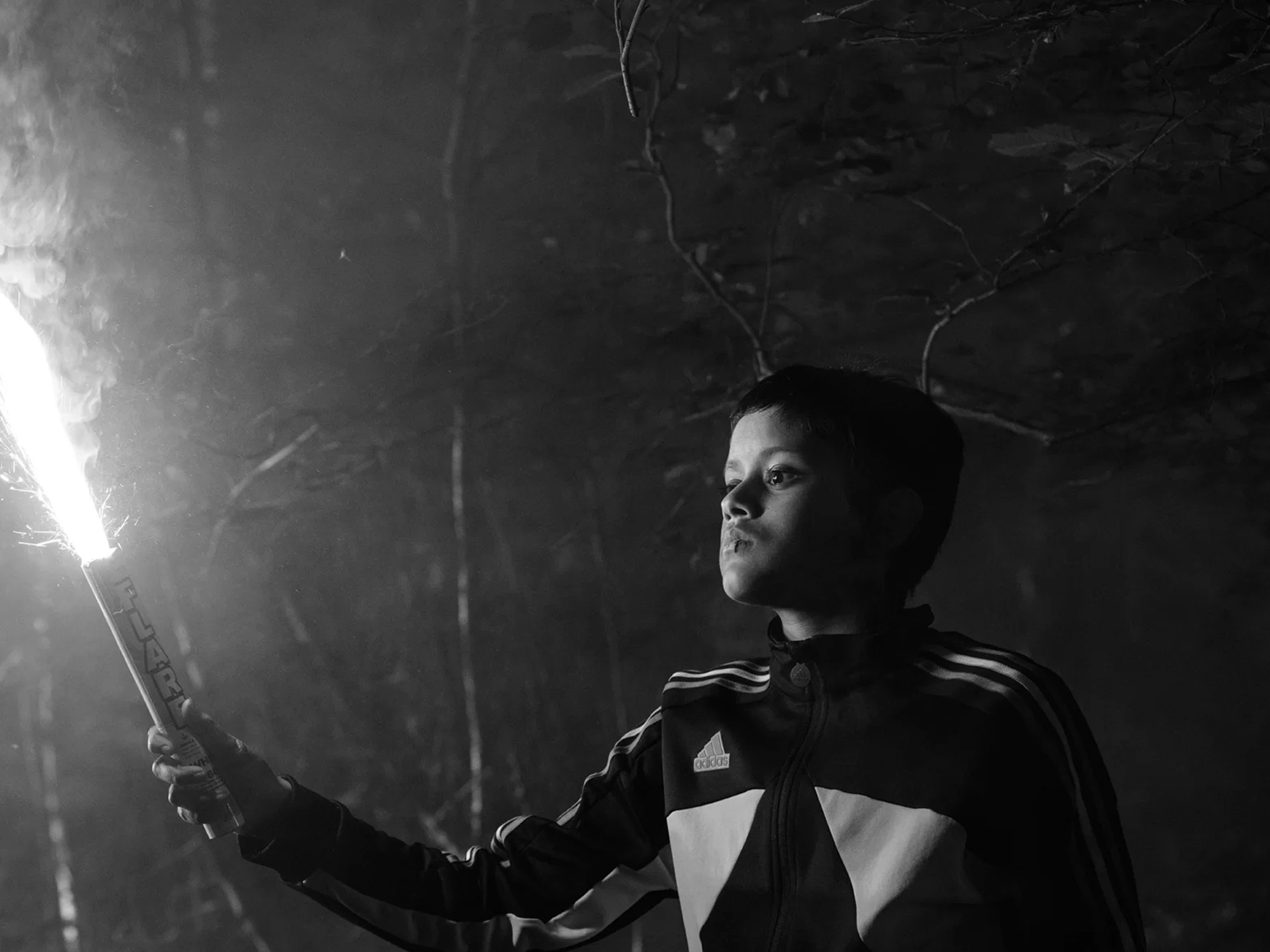 Black and white image of person in dark clothing holding bright light source, standing against textured ceiling background.