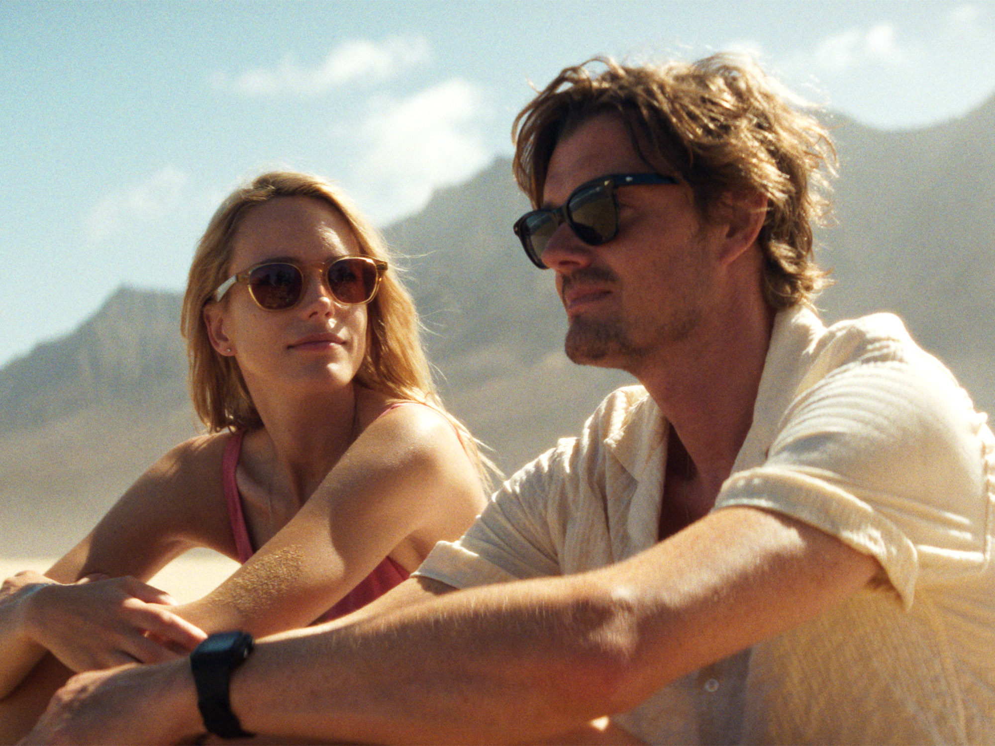 Two people wearing sunglasses sitting together outdoors with mountains and blue sky in background, woman in red bikini, man in white shirt.