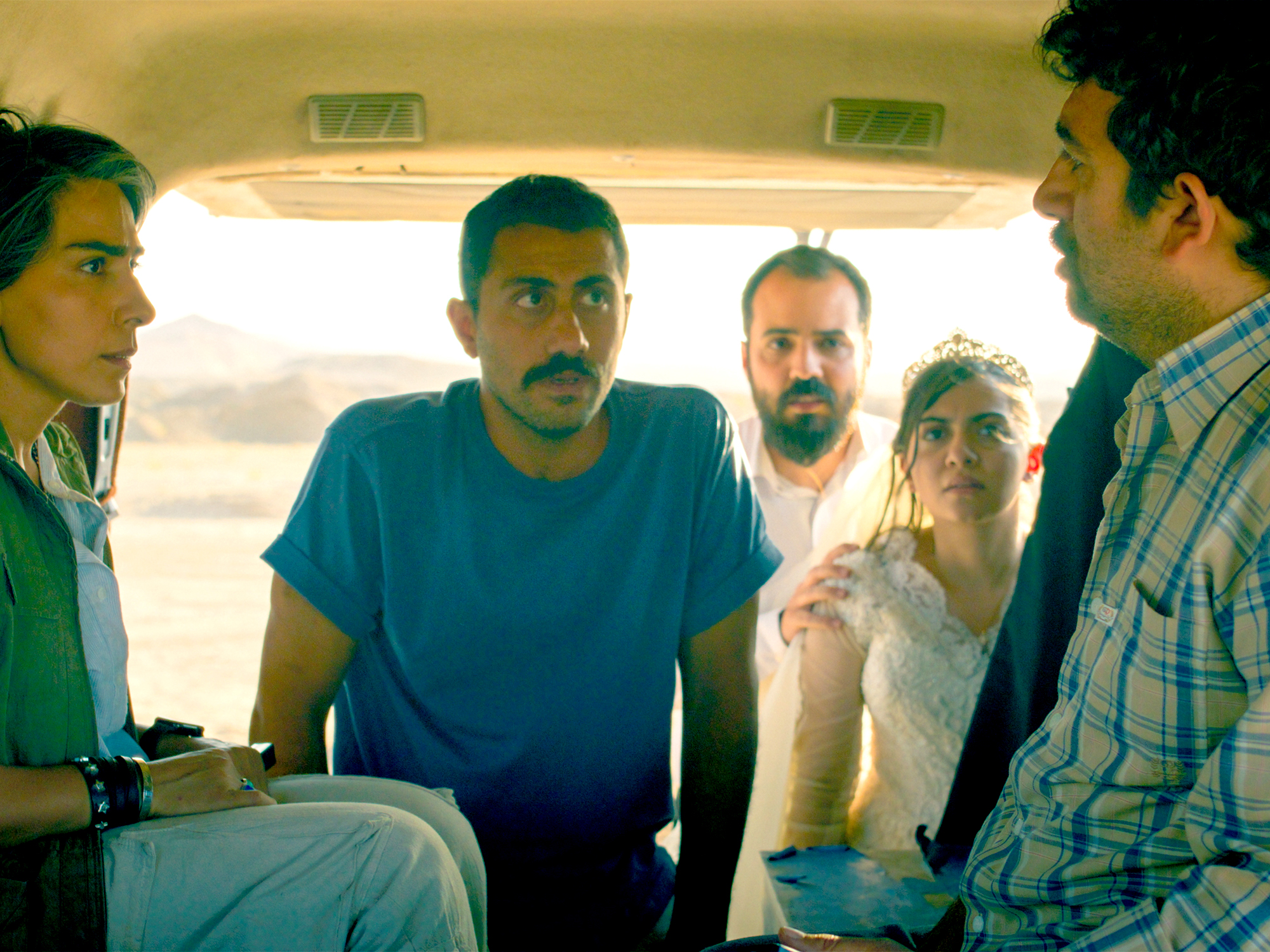 Five people sitting inside a vehicle, with warm yellow lighting from windows behind them creating a bright backdrop against their silhouettes.