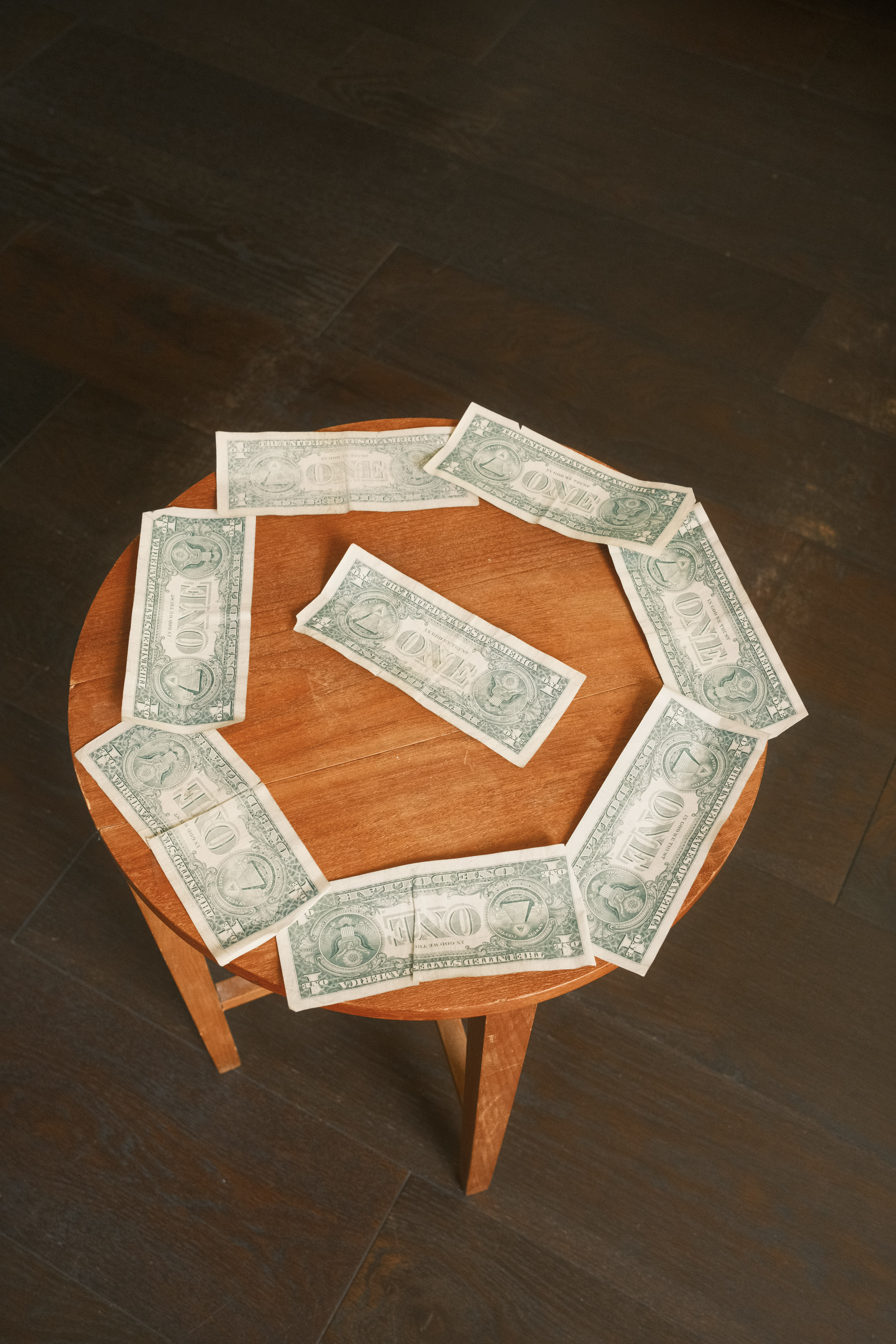 Eight one-dollar bills arranged in a circle on a round wooden table, with one bill placed in the centre, against a dark background.