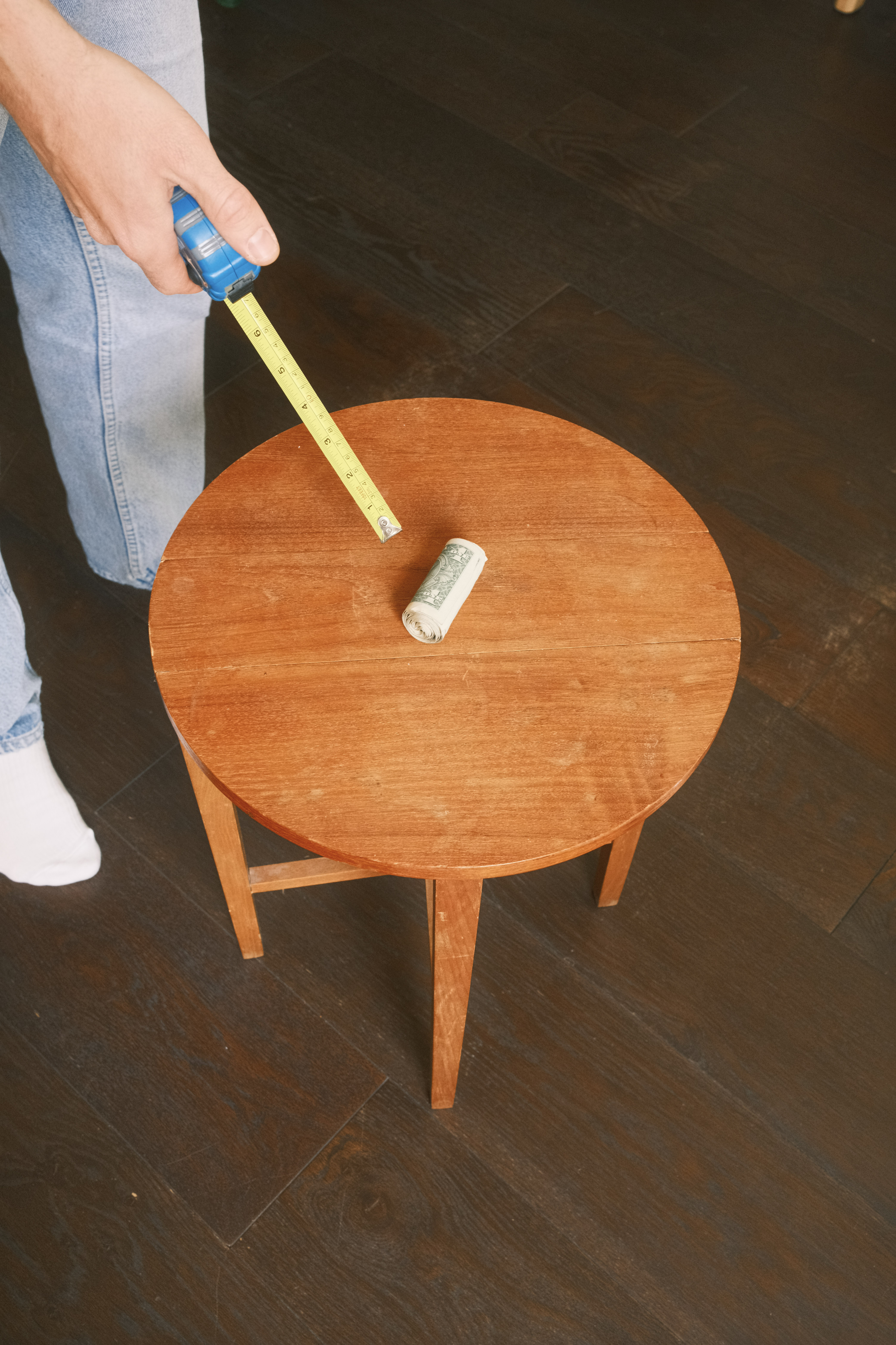 Person holding measuring tape across wooden round table with white cloth on dark tiled floor.