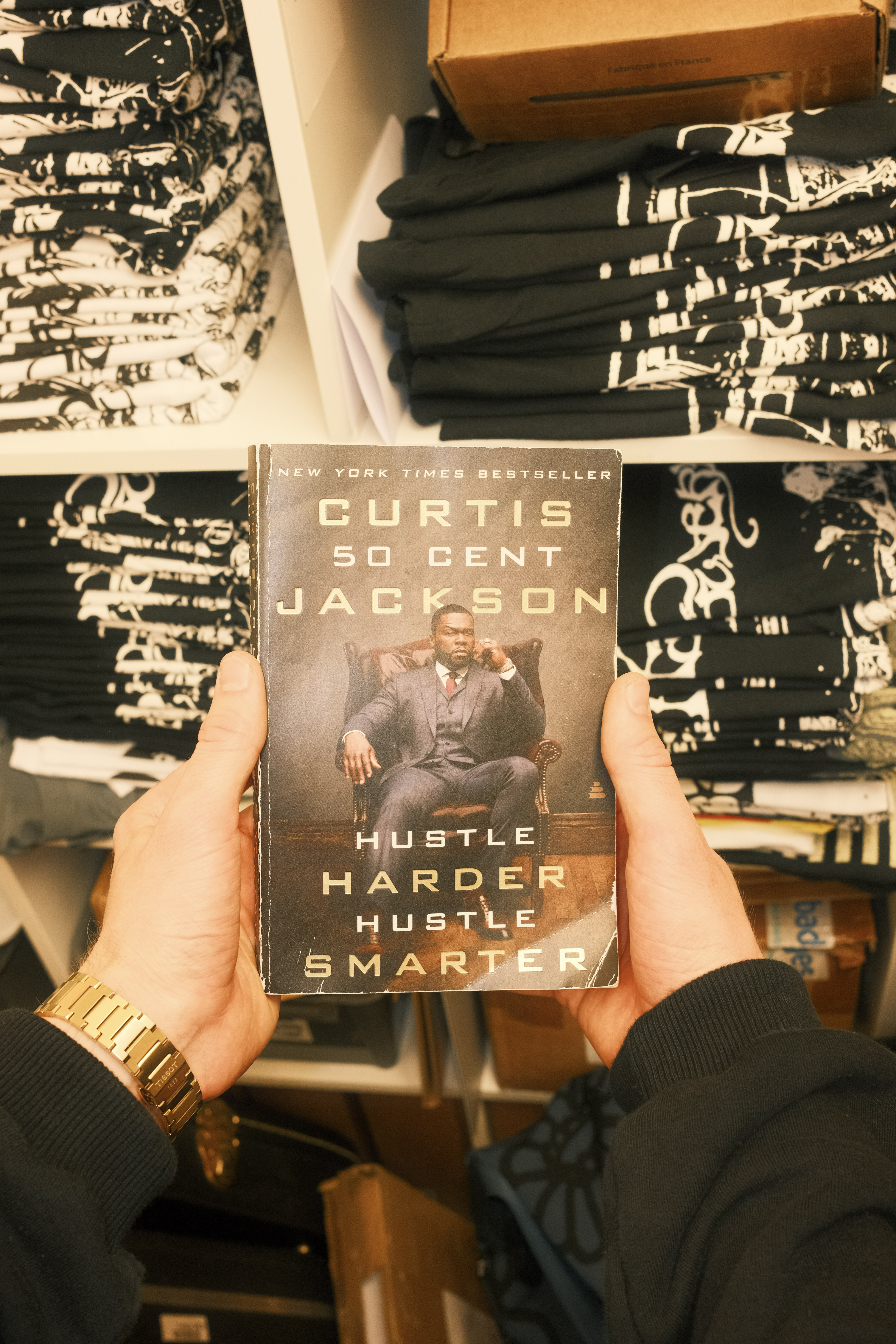 Hands holding "Hustle Harder Hustle Smarter" book by Curtis "50 Cent" Jackson in bookshop, with stacks of books in background.