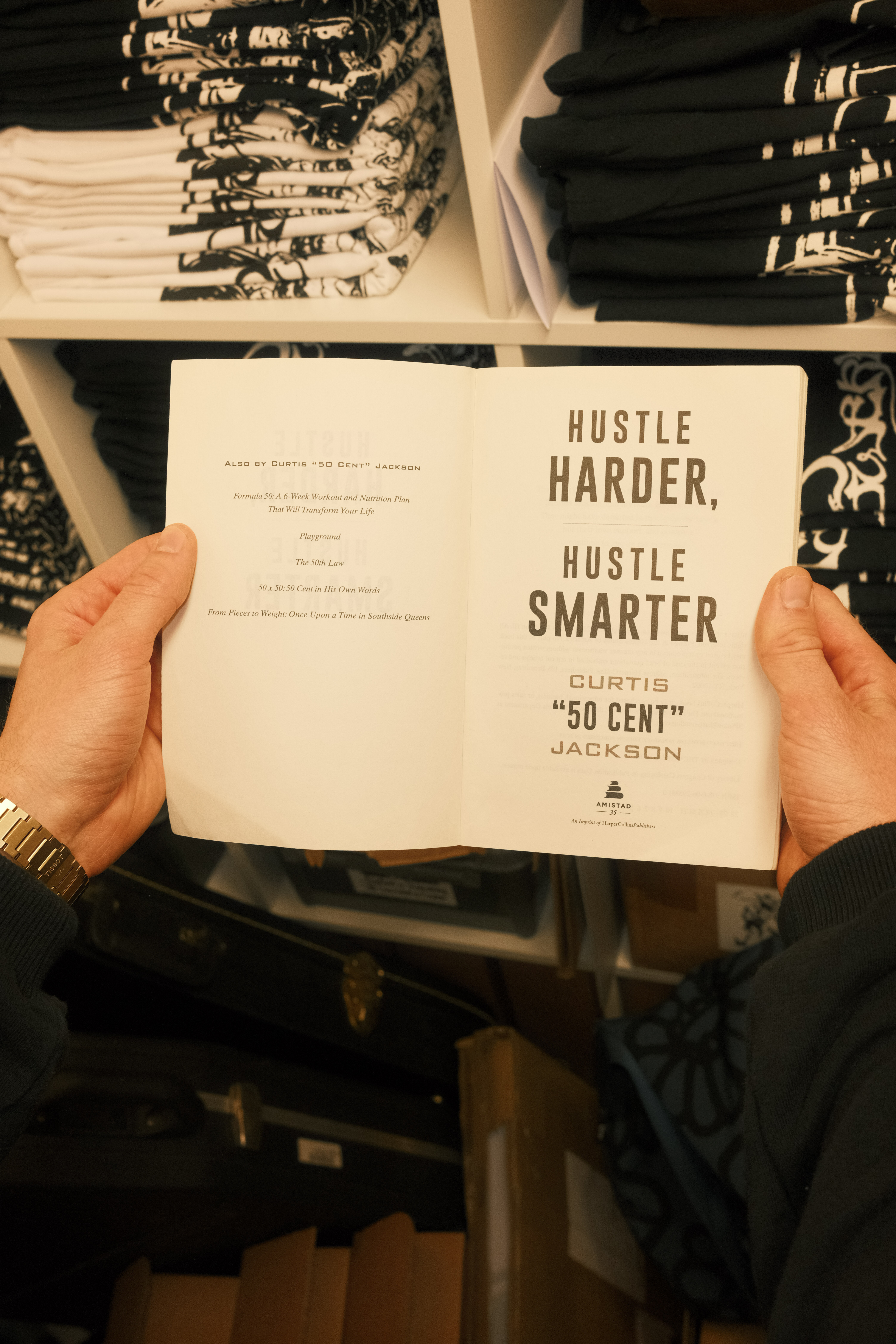 Hands holding open book titled "Hustle Harder, Hustle Smarter" by Curtis "50 Cent" Jackson in clothing shop with racks of garments.