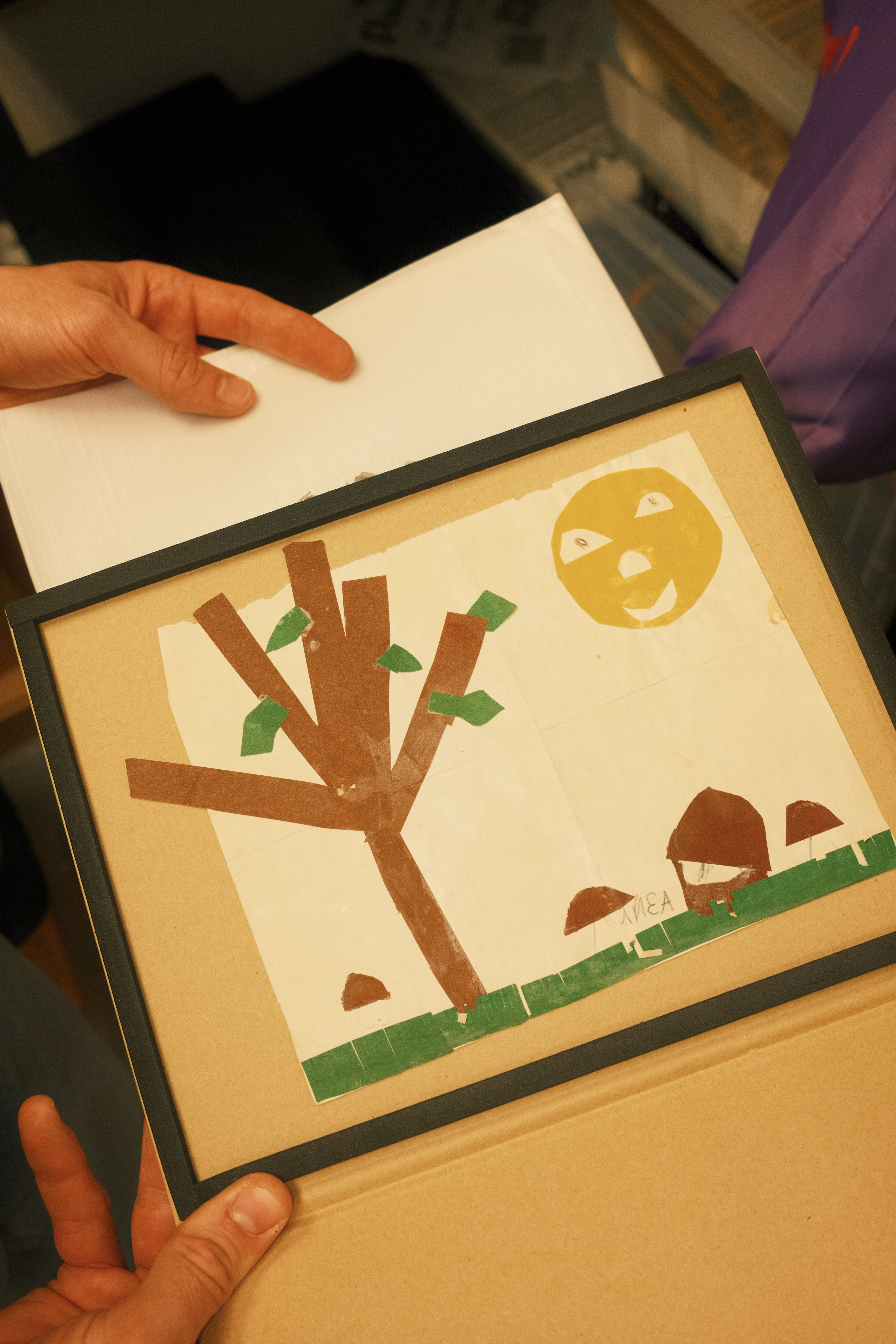 Hands holding children's artwork with brown paper tree, green leaves, yellow sun, and brown mushrooms on cream background.