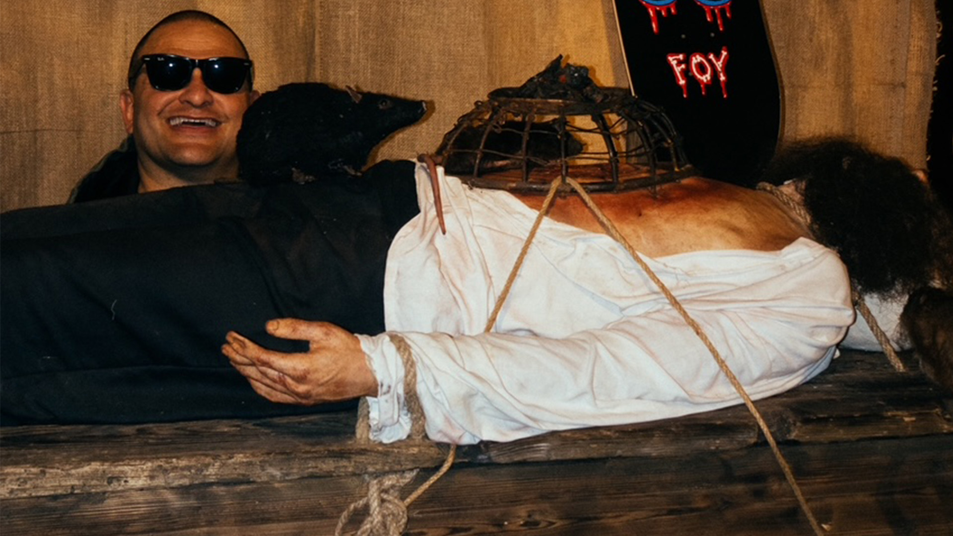 Man in dark clothing and sunglasses sits beside figure wrapped in white cloth with metal cage-like headpiece against brick wall.