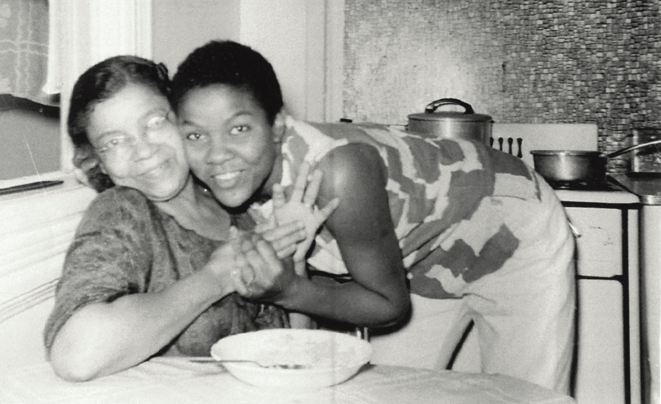 Black and white image of two smiling women embracing at a kitchen table, with cooking pots visible on the hob behind them.