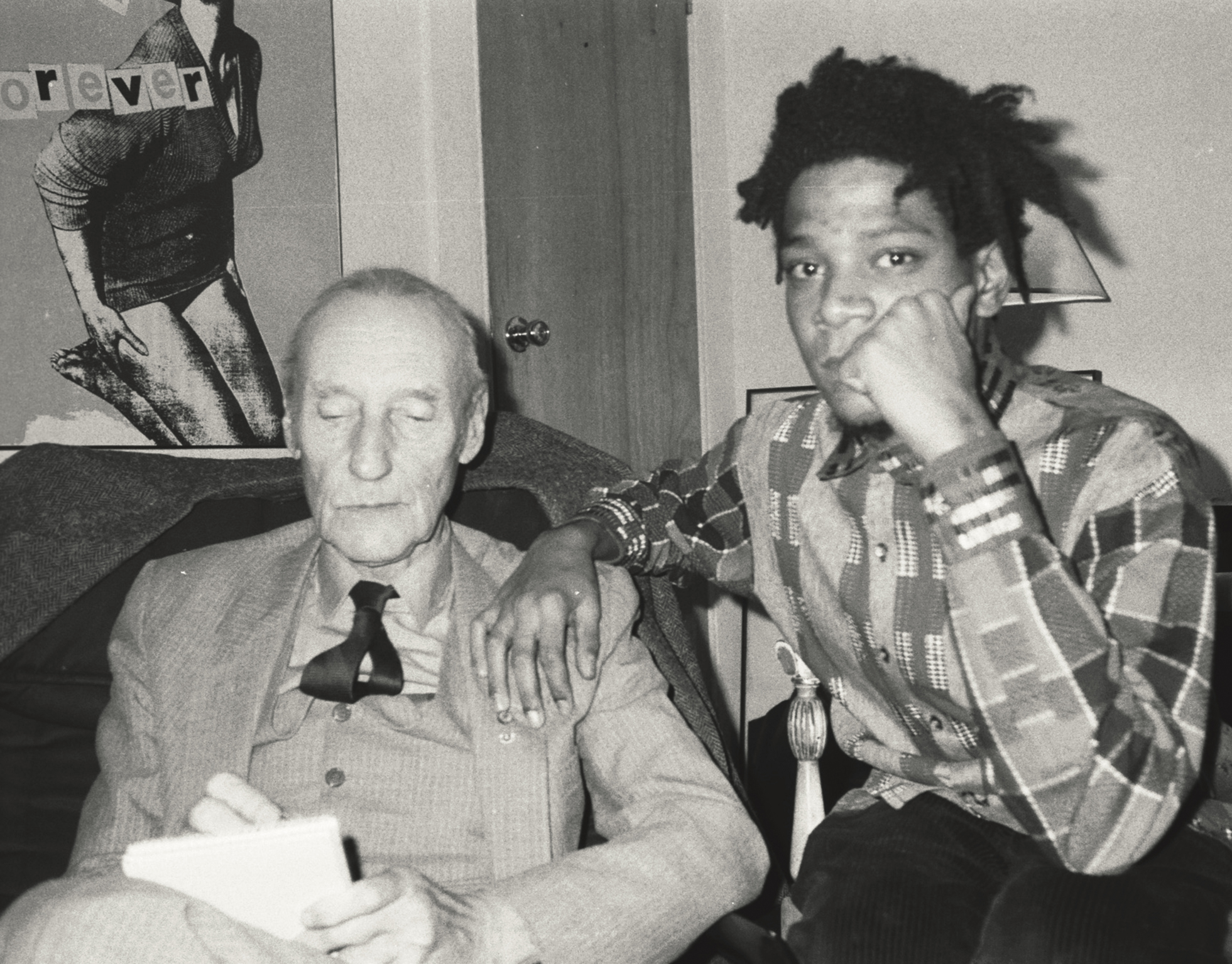 Elderly white man in suit and tie sits beside young Black man with dreadlocks wearing plaid shirt, both on sofa in black and white image.
