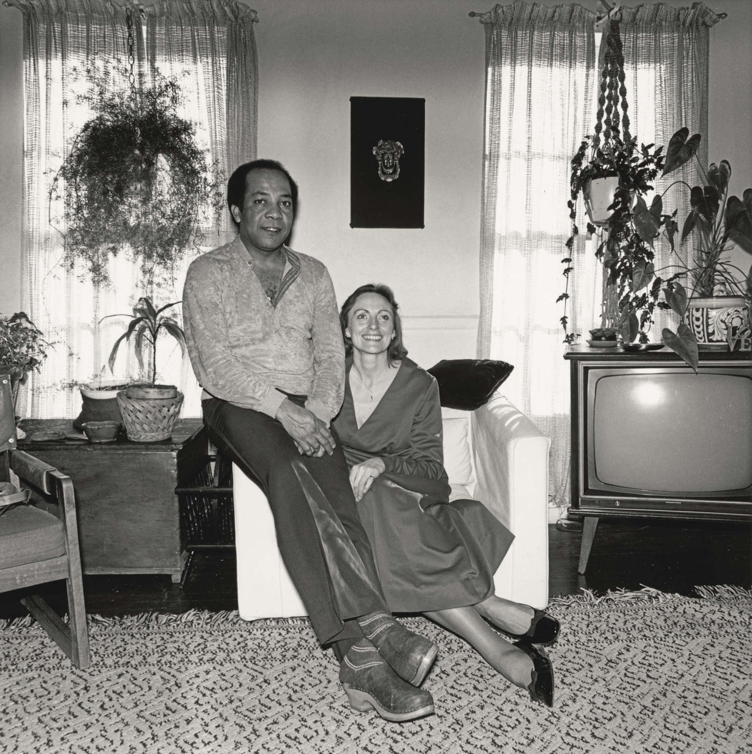 Man in jumper standing beside woman seated in chair in 1970s living room with television, plants, and patterned rug.