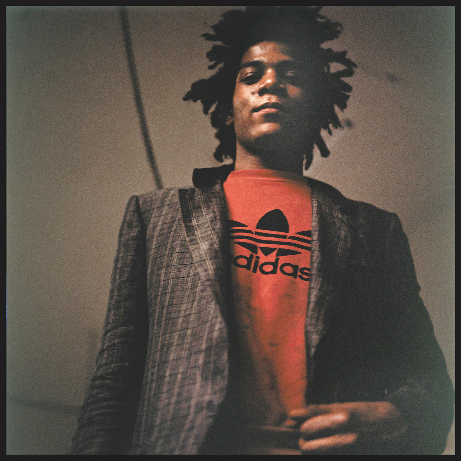Man with dreadlocks wearing red Adidas t-shirt and grey blazer, looking upwards against neutral background.
