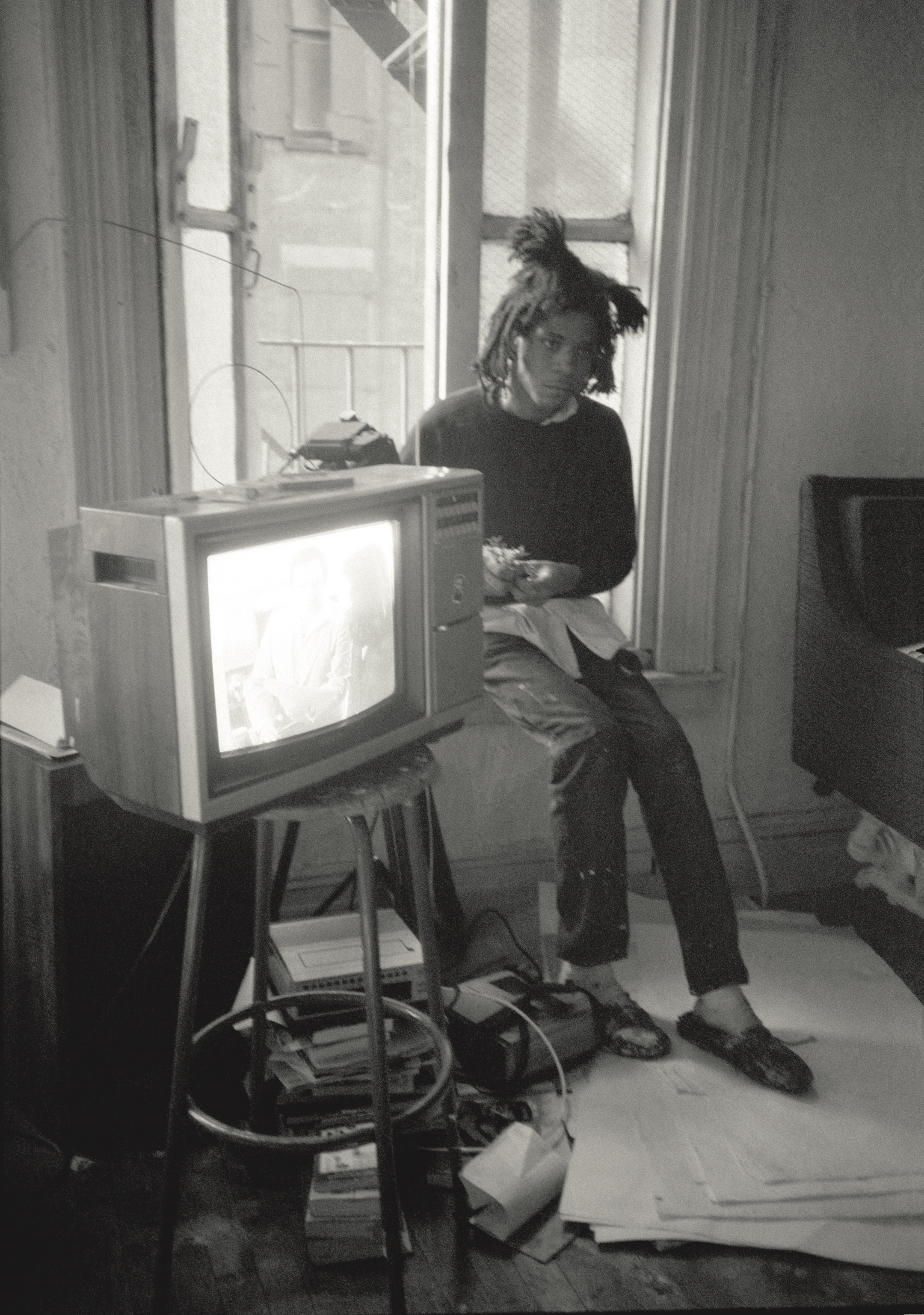 Person with dreadlocks sitting on stool next to vintage television on stand, holding bowl, black and white photograph