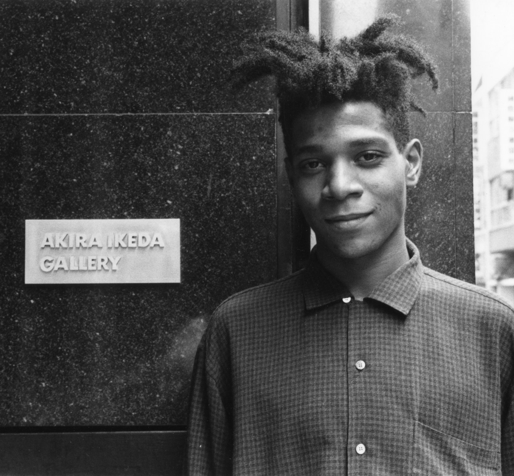 Young Black man with locs in checked shirt stands beside dark wall with "AKIRA IKEDA GALLERY" nameplate in black and white portrait.