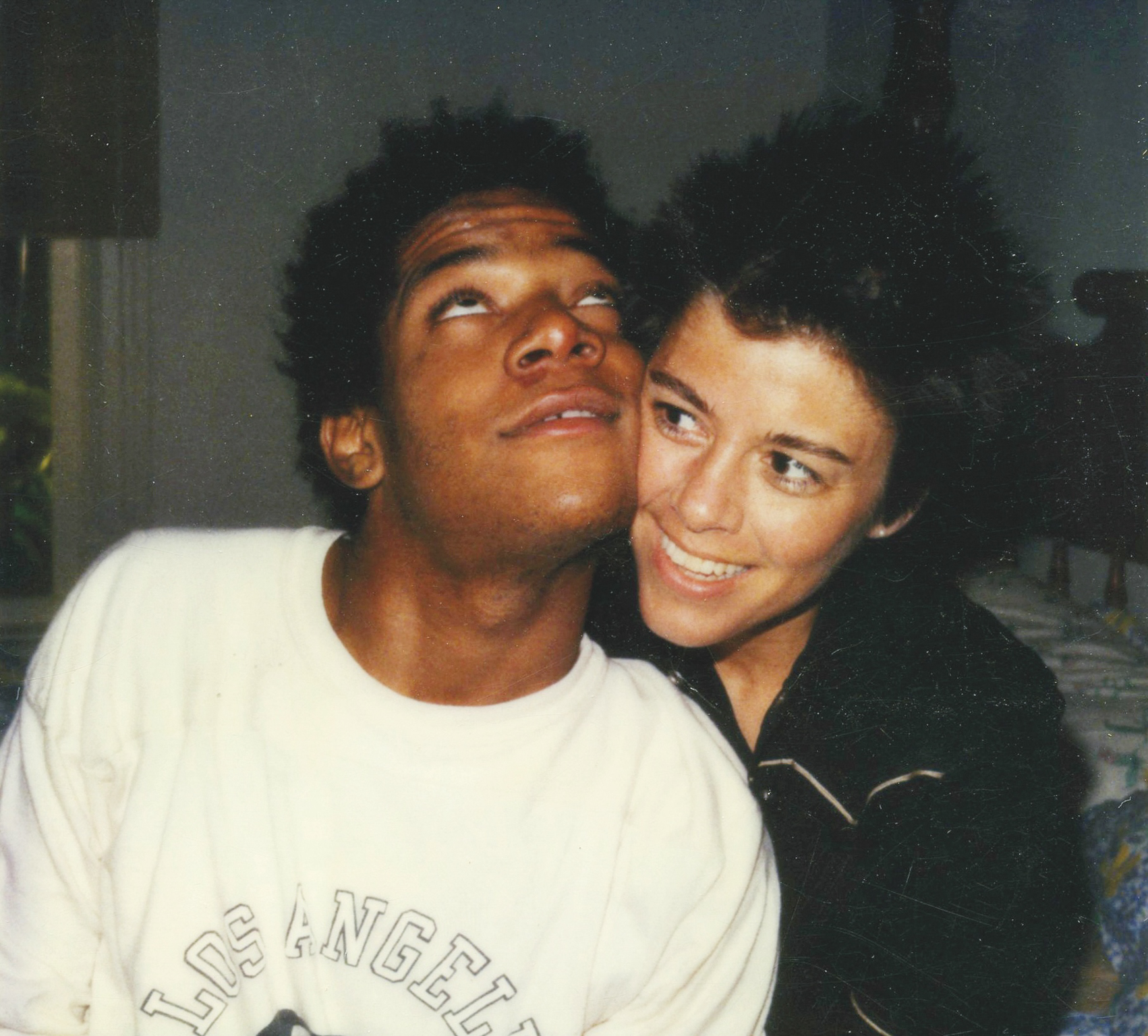 Young man in white Lakers t-shirt with woman in black top embracing closely, both smiling in indoor setting with dim lighting.