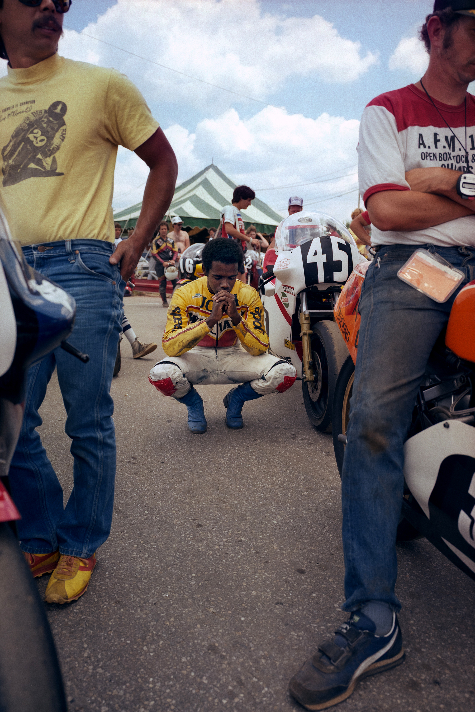 Racing driver in yellow suit crouched between go-karts numbered 43 and 19, surrounded by spectators in casual clothing at outdoor karting event.