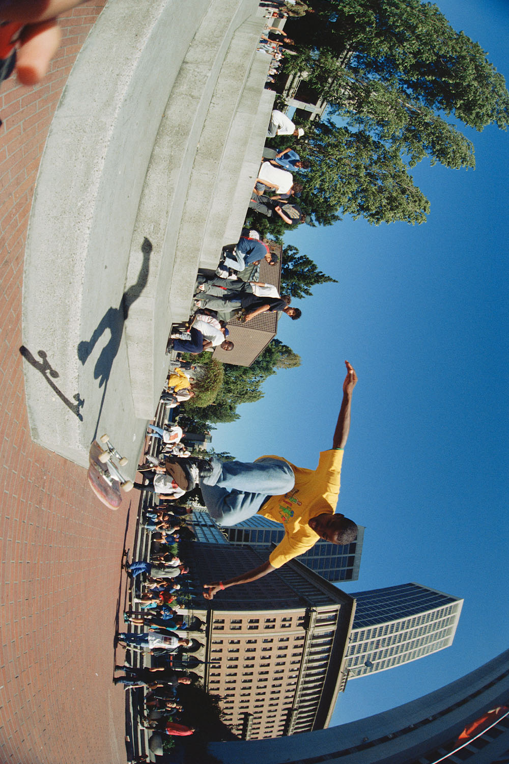 Rotated image: person in yellow top mid-jump on pavement, tall buildings and trees in background, clear blue sky, market stalls visible.