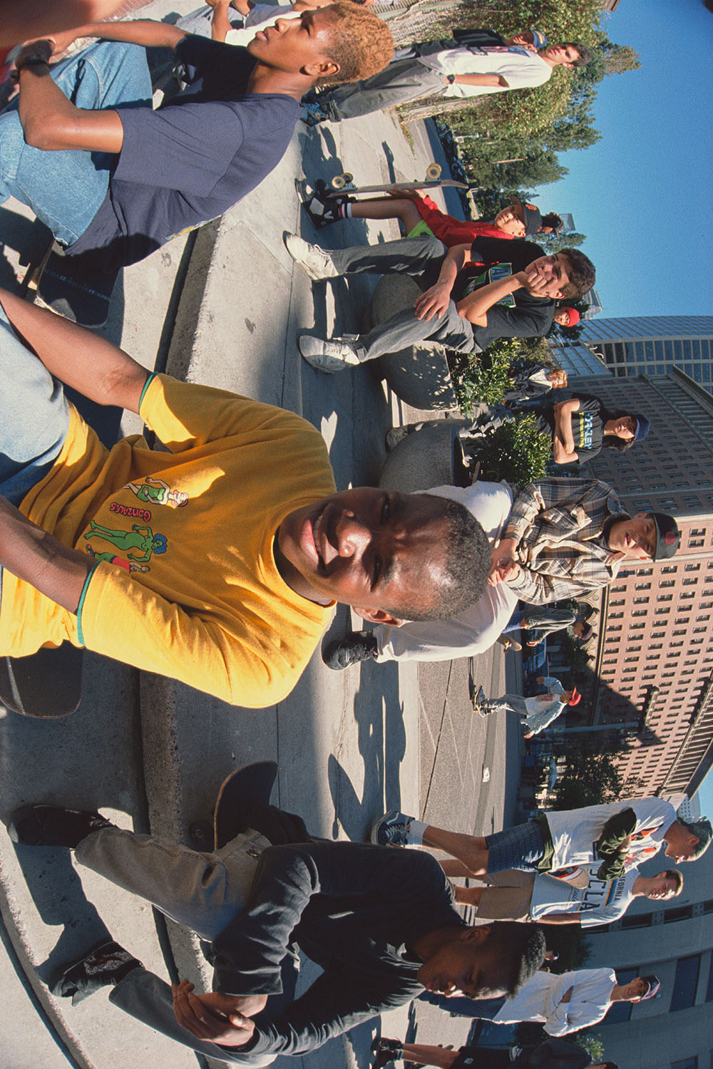 Man in yellow shirt speaking at microphone to crowd gathered on street with buildings and trees in background.
