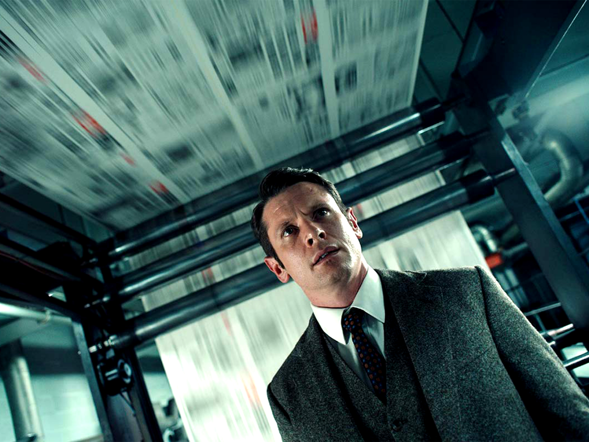 Man in dark suit and tie inside moving vehicle with blurred motion lines and teal-green tinted lighting creating dynamic streaked effect.