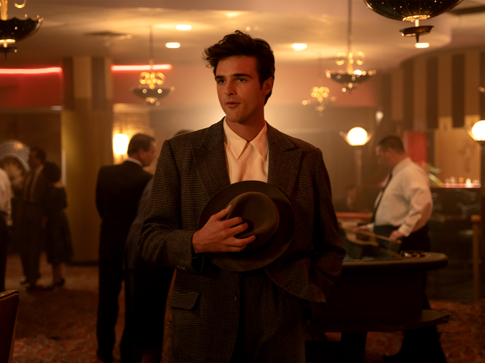 Man in dark suit holding helmet in upscale restaurant with warm lighting and other diners in background.