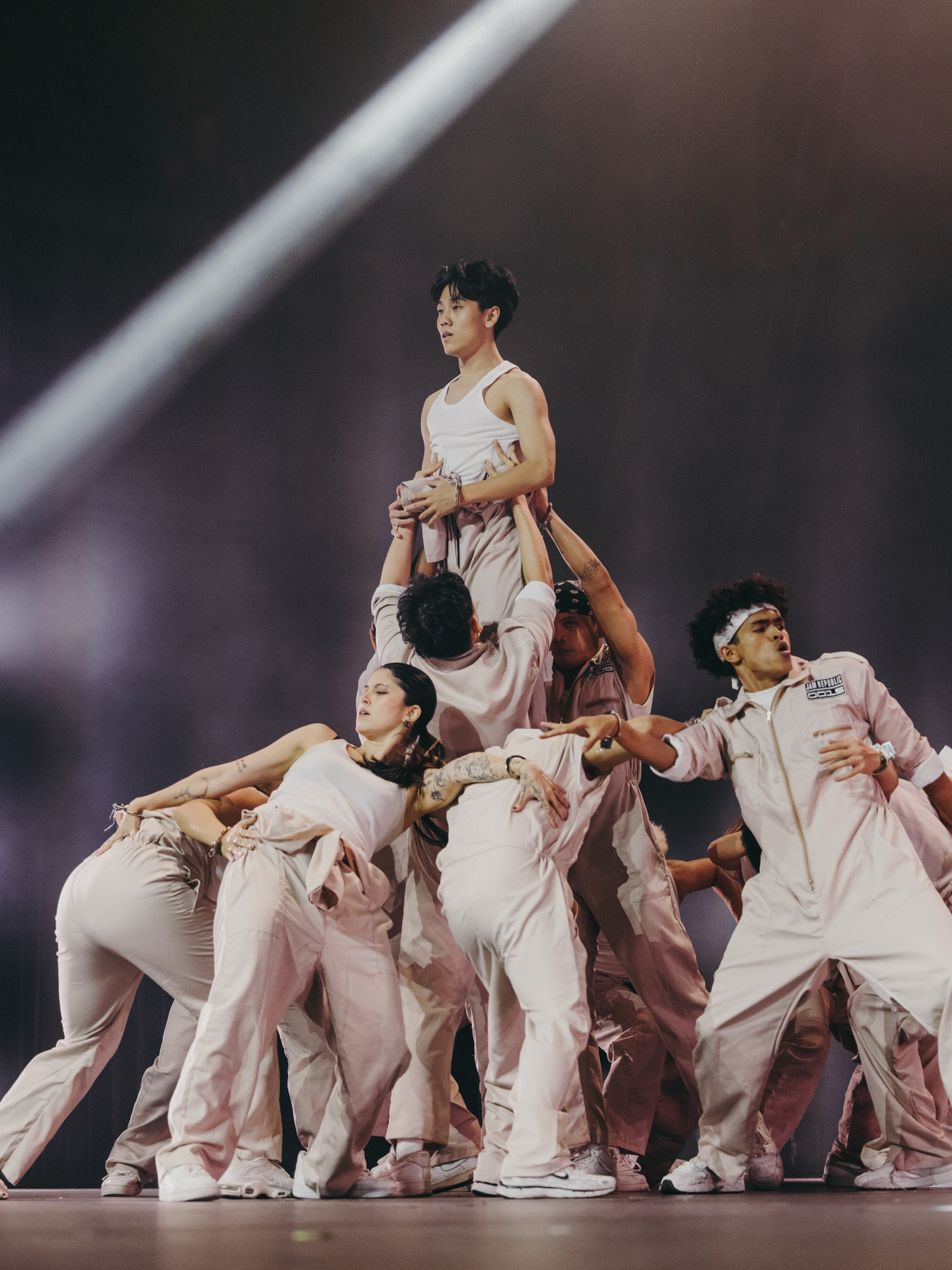 Group of performers in white tracksuits forming human pyramid on stage with dramatic lighting beams in background.