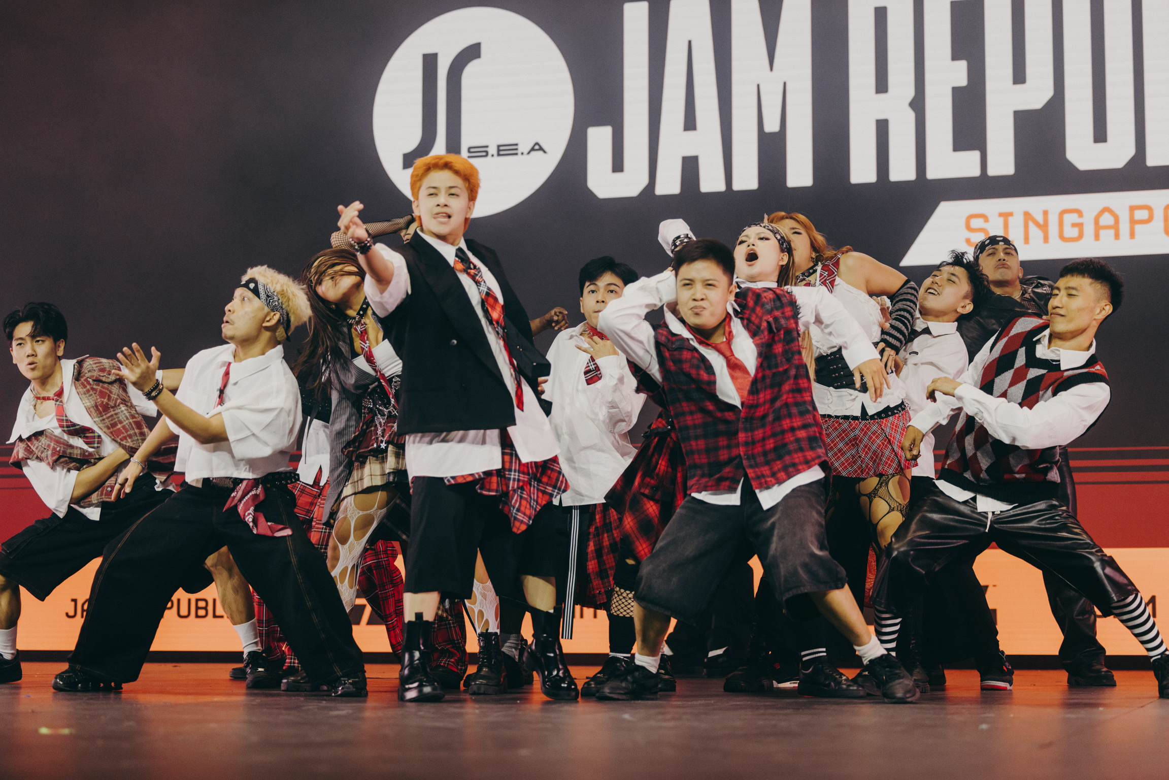 Performance group on stage wearing black suits and red plaid outfits, dancing with raised arms against backdrop reading "JAM REPUB".