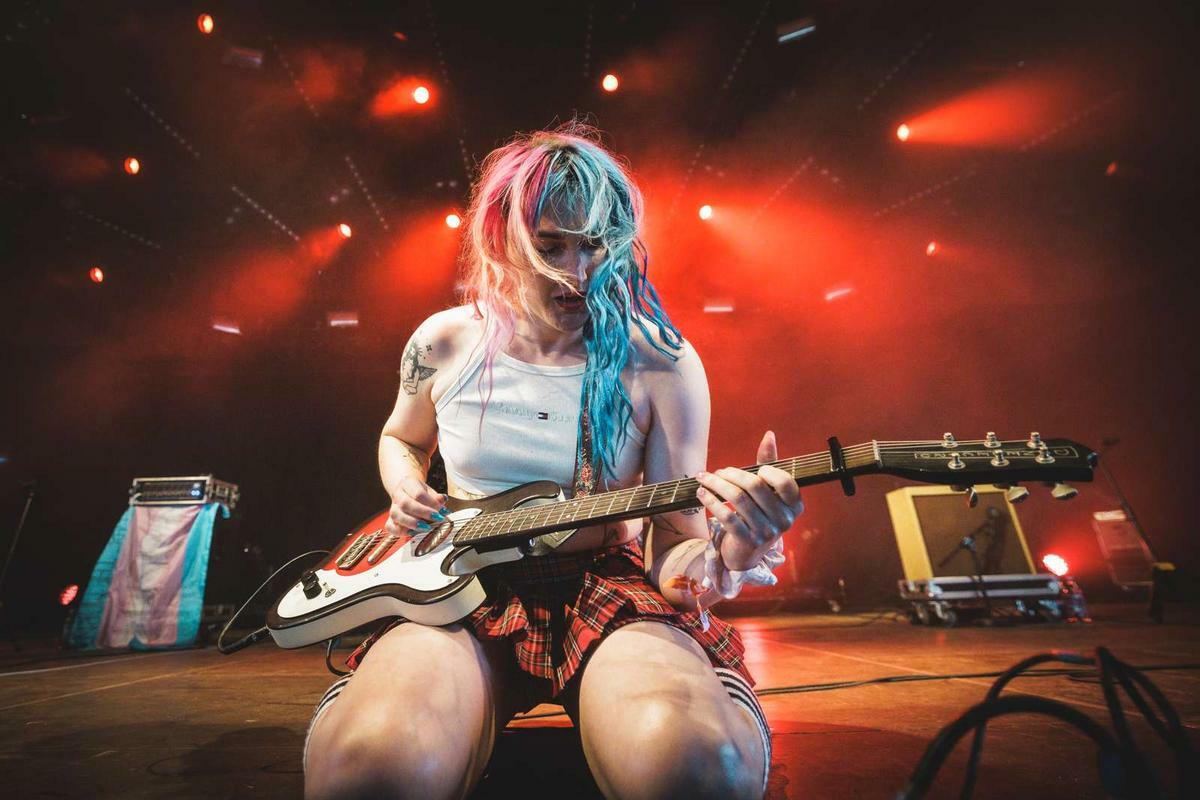 Performer with pink and blue hair sits on stage playing white electric guitar, wearing white top and plaid skirt under red stage lighting.