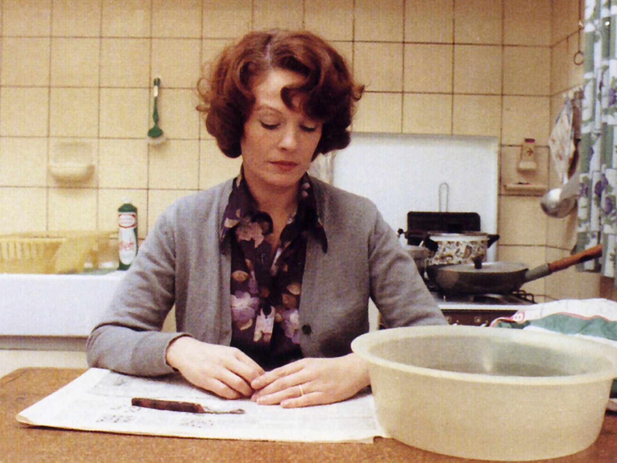 A woman with curly red hair wearing a grey cardigan and floral scarf, sitting at a kitchen table, looking focused as she writes on a piece of paper.