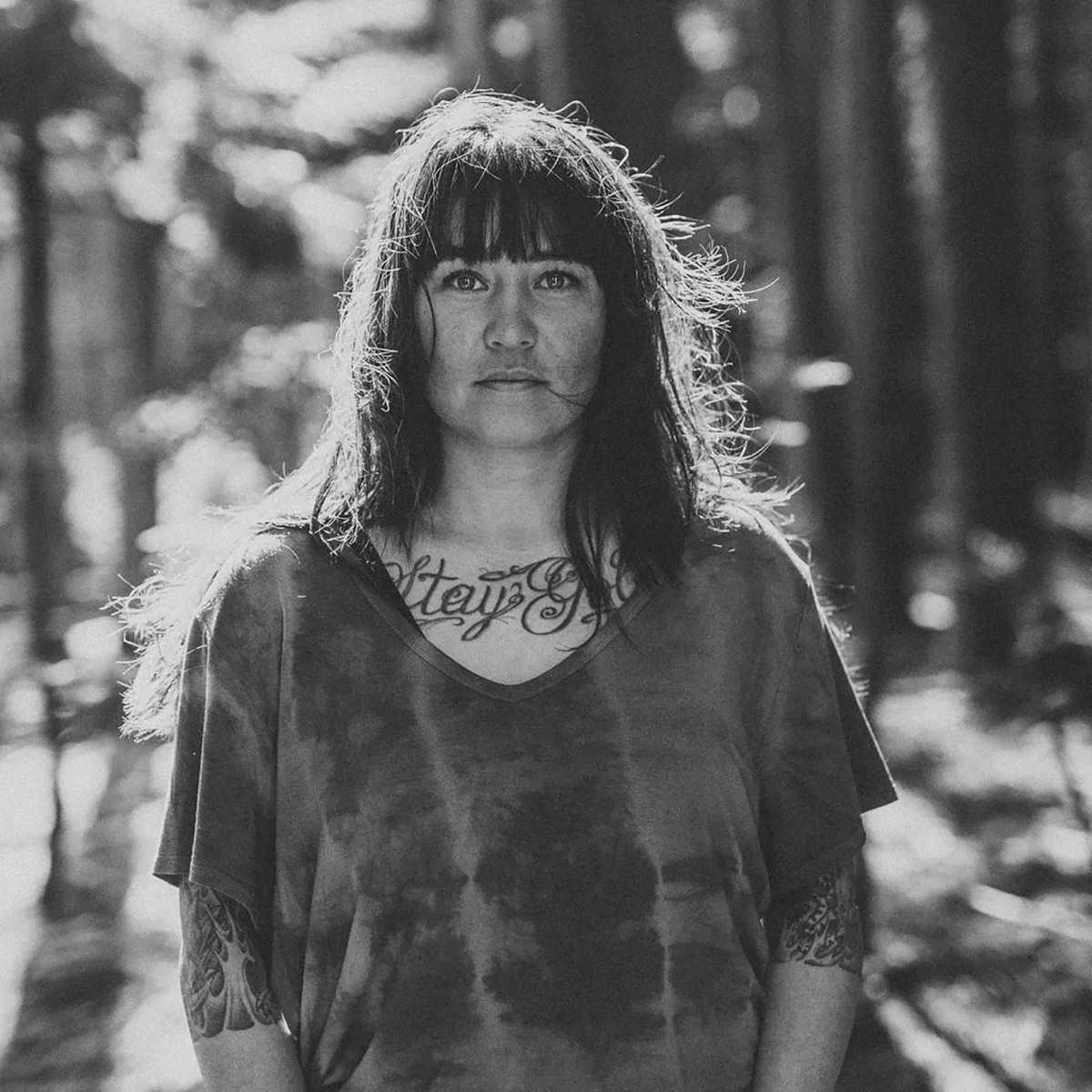 Black and white portrait of woman with fringe and shoulder-length hair, wearing v-neck top, chest tattoo visible, blurred forest background.