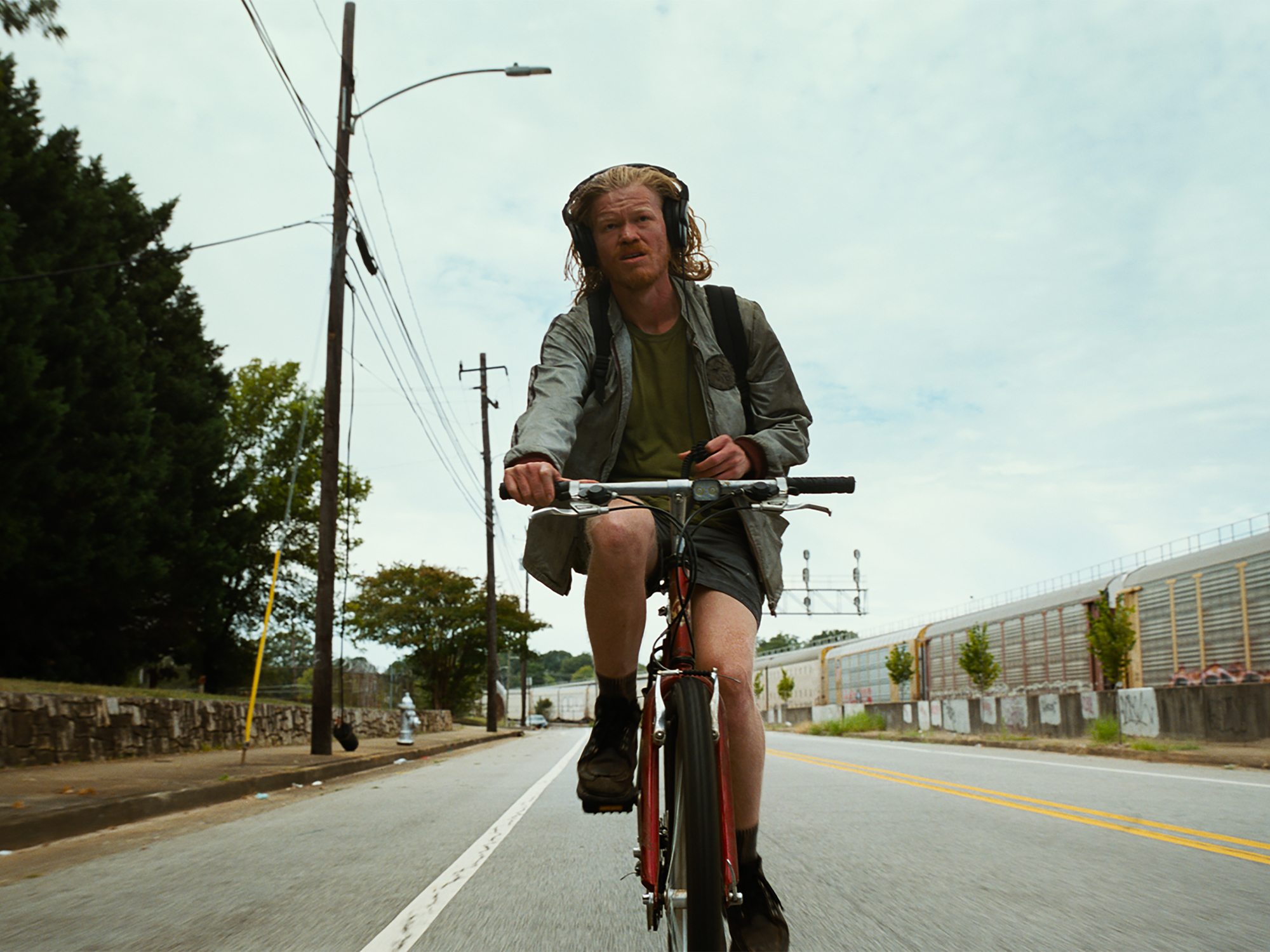 Man cycling on empty road wearing grey jacket and green top, with power lines and trees visible alongside street.