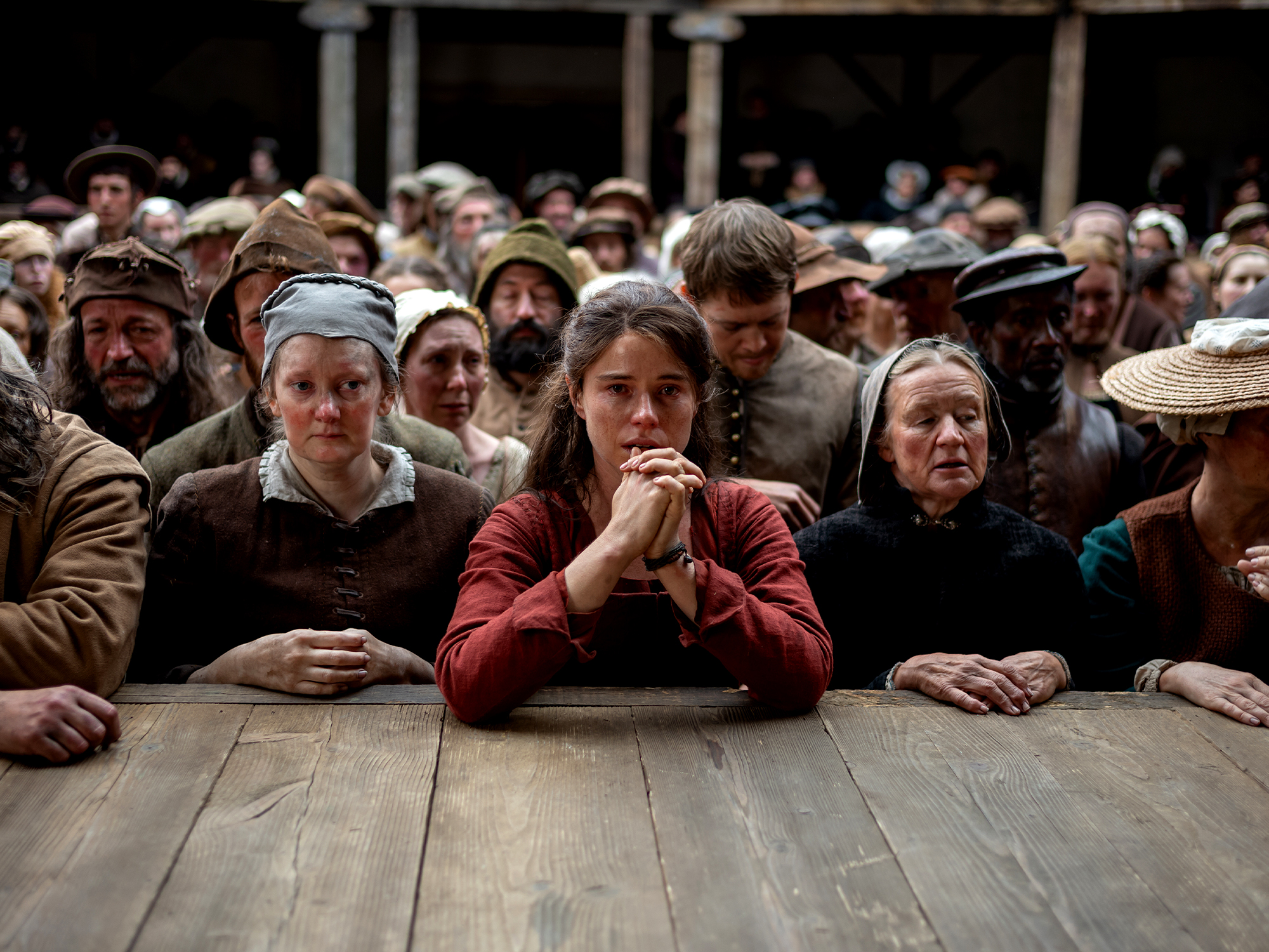Crowd of people in period dress gathered around wooden table, woman in red at centre with hands clasped, others wearing caps and bonnets.