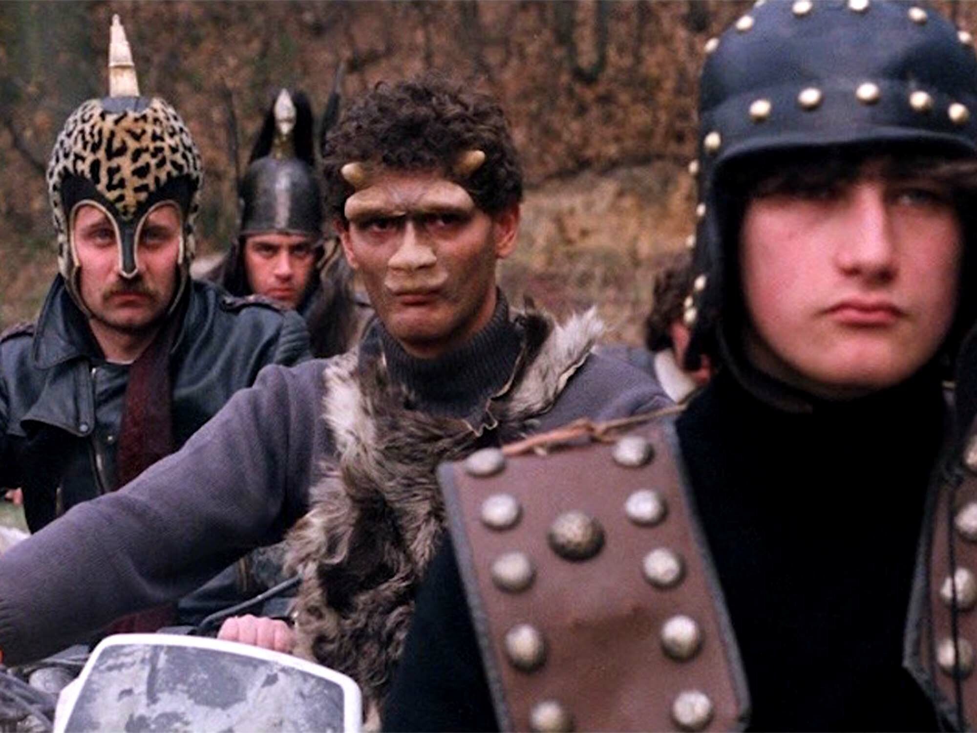 Four individuals wearing post-apocalyptic costumes including helmets, goggles, and fur-trimmed clothing, holding various props like shields and weapons.