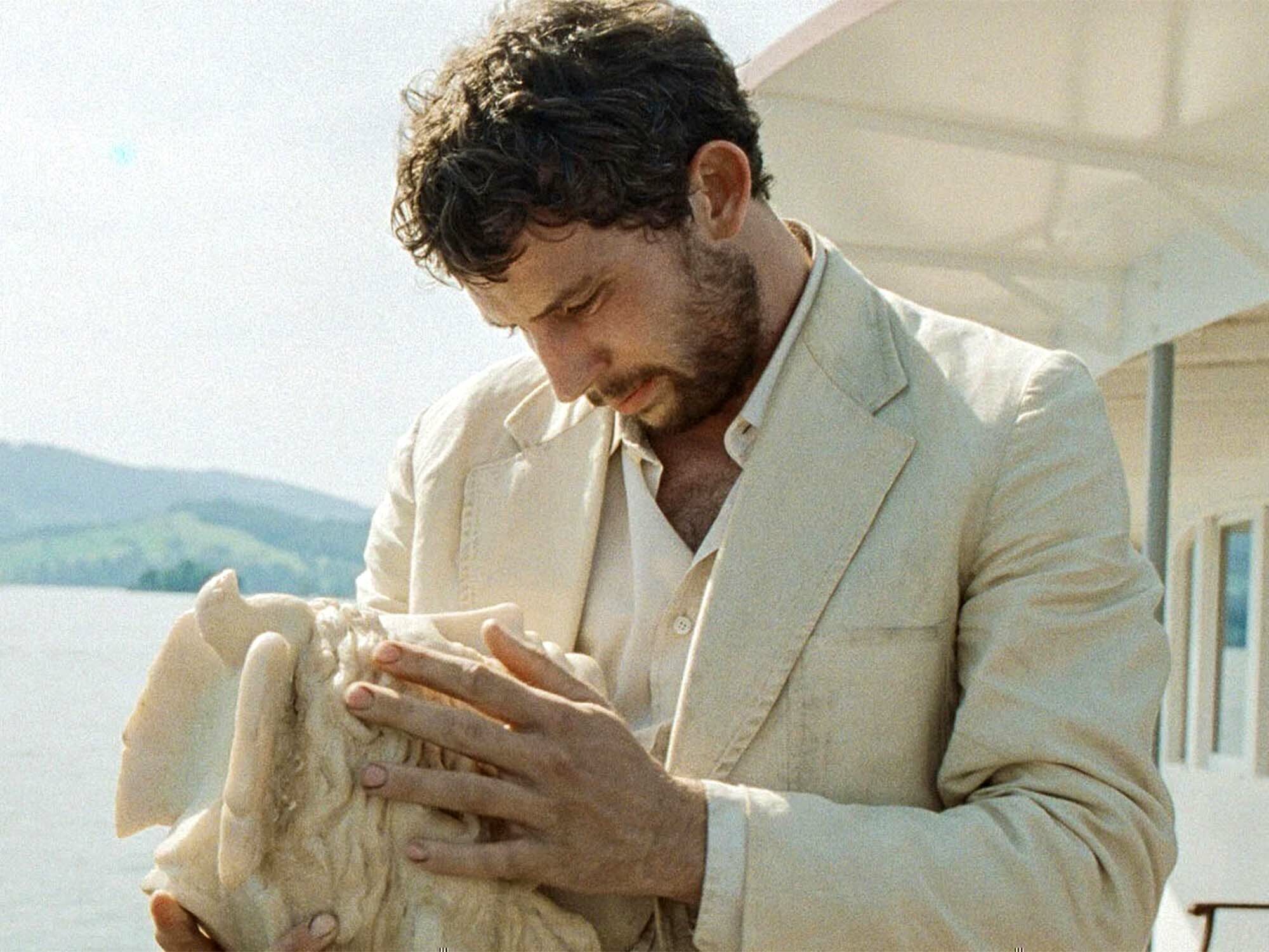 A man with curly hair and a beard, wearing a light-coloured jacket, sitting on a bench and holding a small white dog.