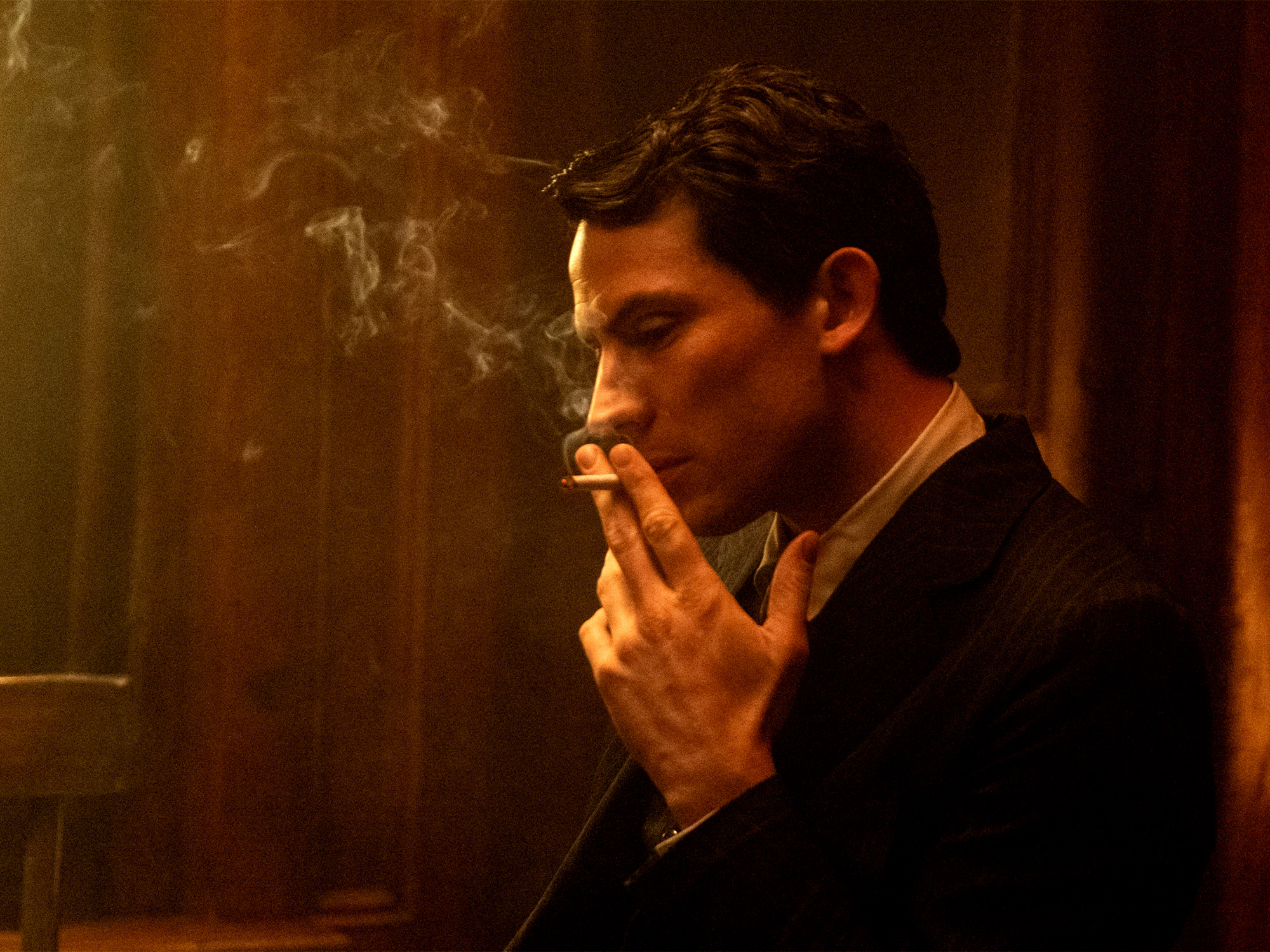 Man in dark suit smoking cigarette in dimly lit room with warm golden lighting and smoke visible in air.