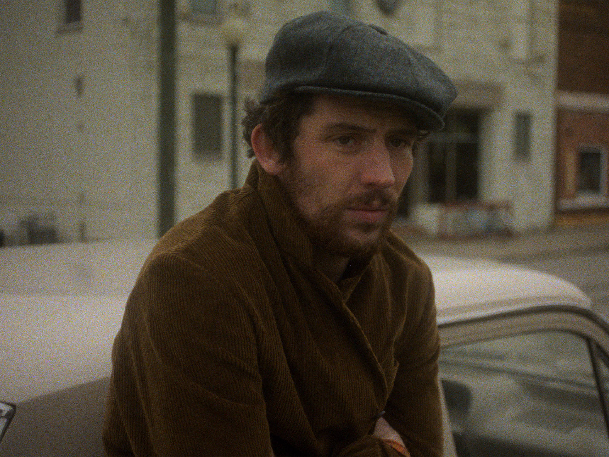 Man in a flat cap, wearing a brown coat, standing in a snowy street next to a parked car.