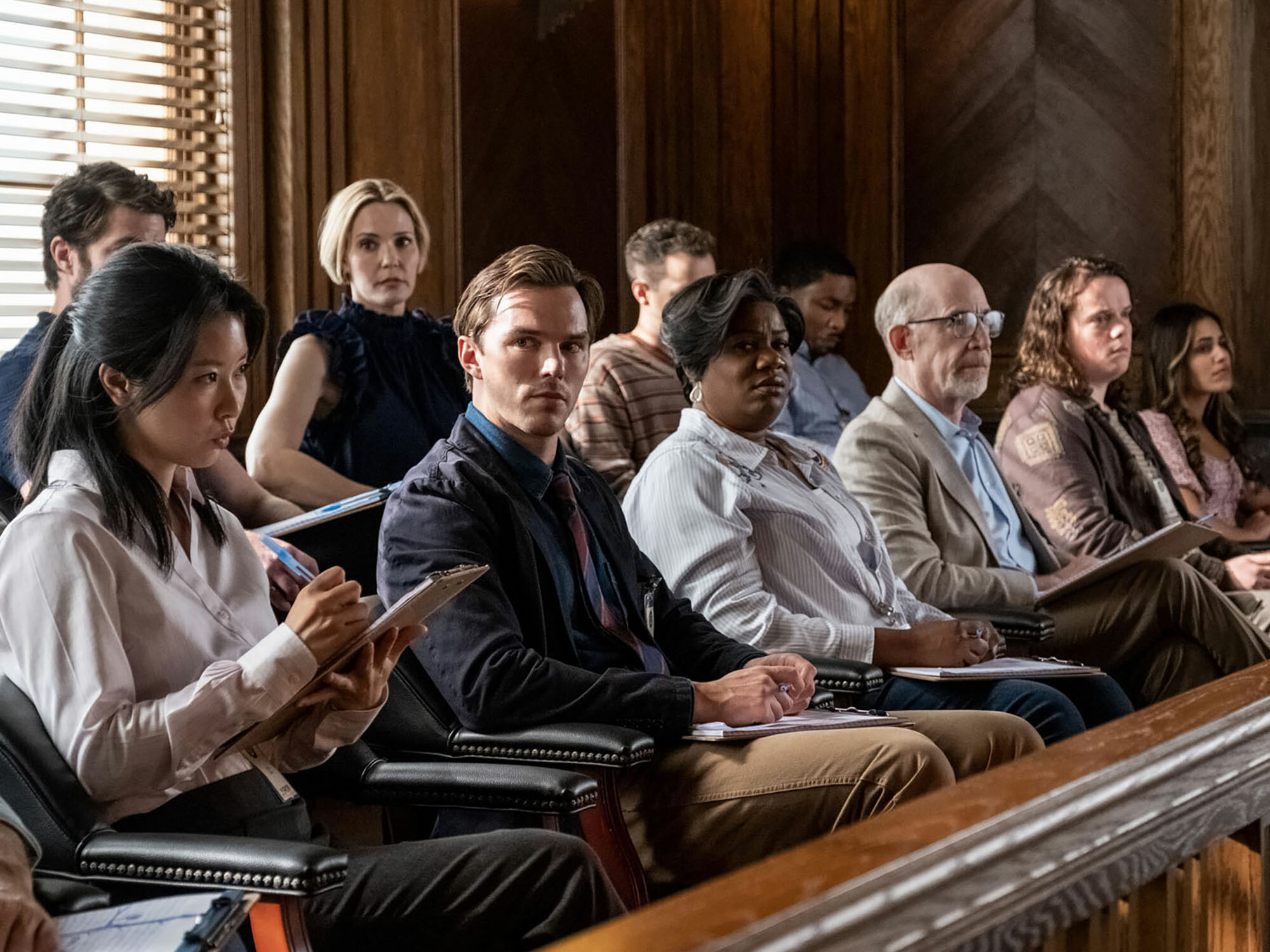 Group of people sitting in courtroom, various ages and ethnicities, some using electronic devices.