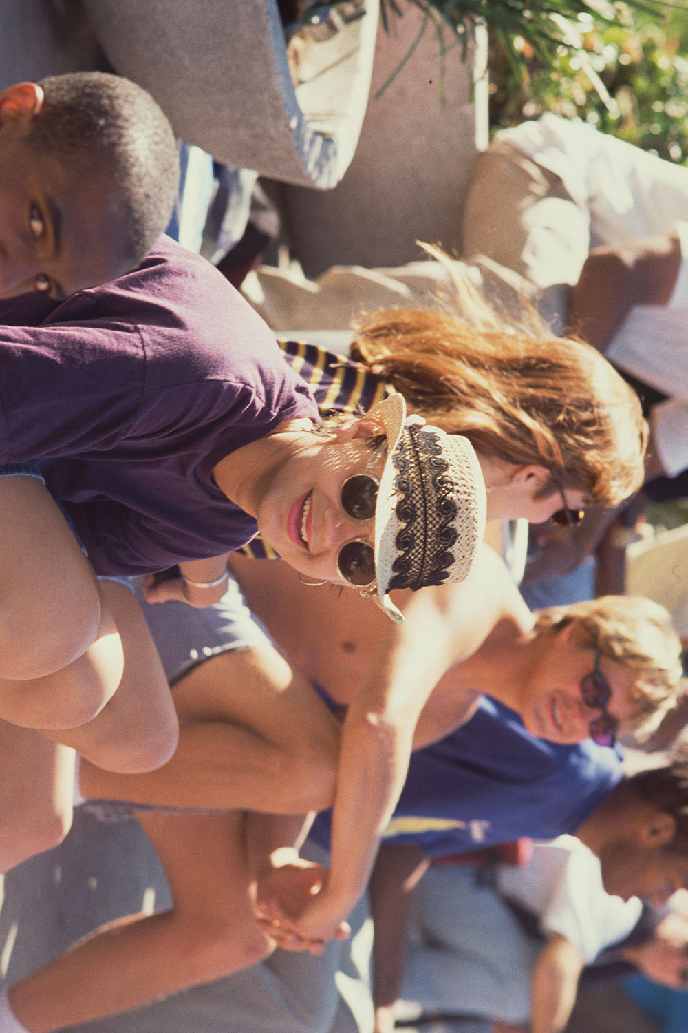 Woman with sunglasses and headscarf smiling outdoors in bright sunlight with other people partially visible in background.