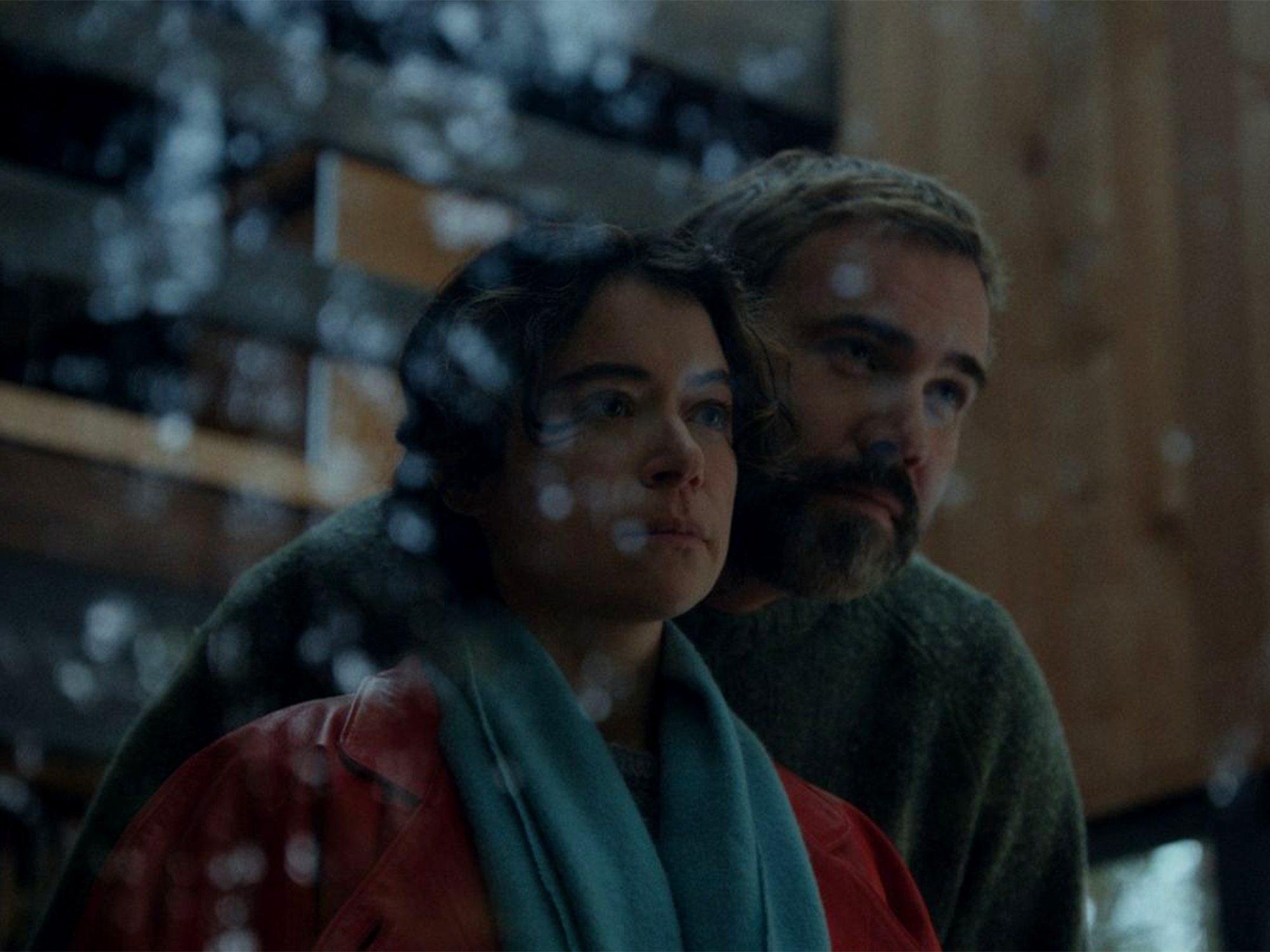 Man with beard embracing woman from behind, both wearing colourful clothing, dimly lit indoor setting with blurred background.