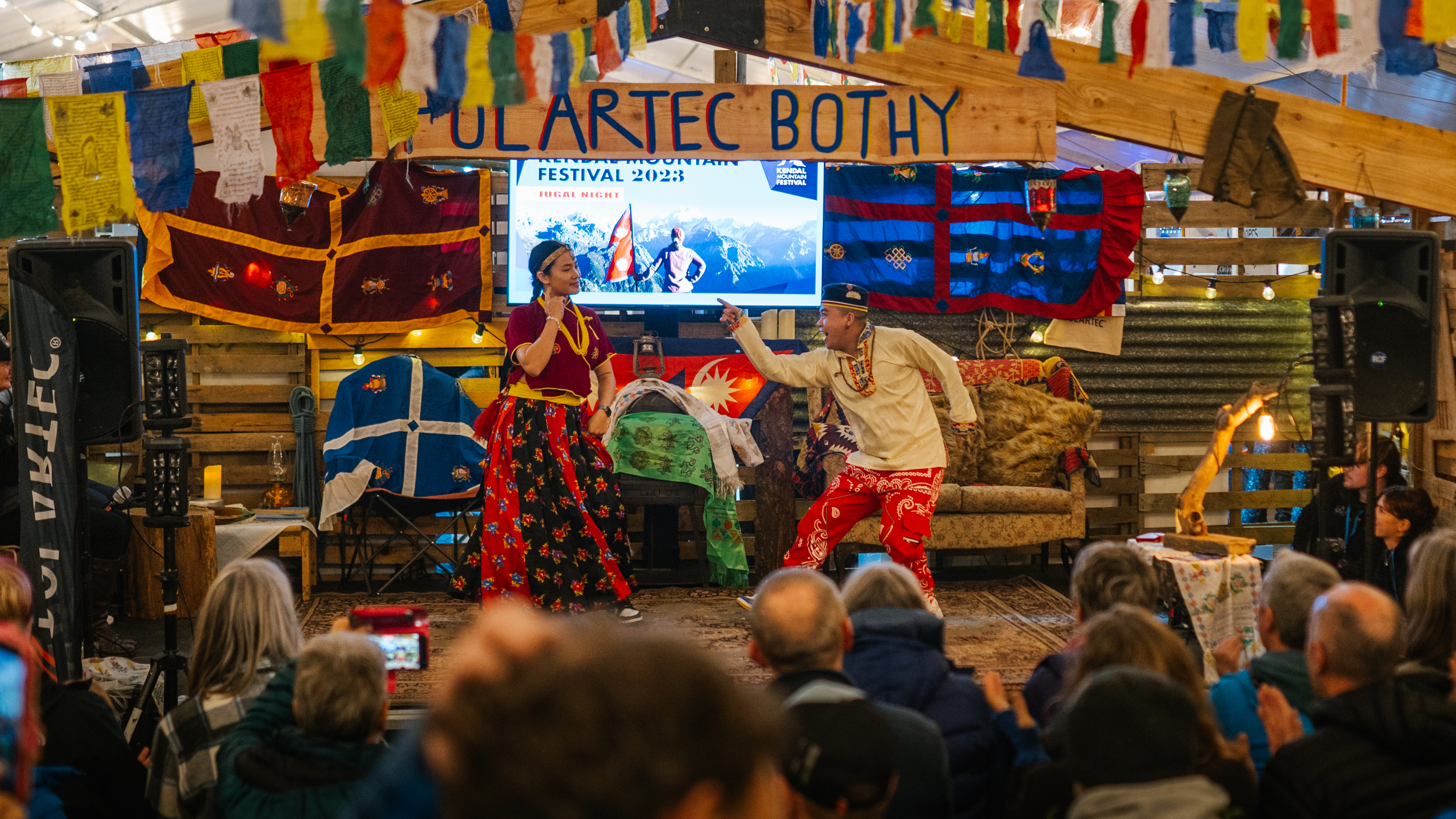 Two performers on wooden stage with colourful prayer flags overhead. Audience seated on floor watching. "Dartec Bothy" sign visible.