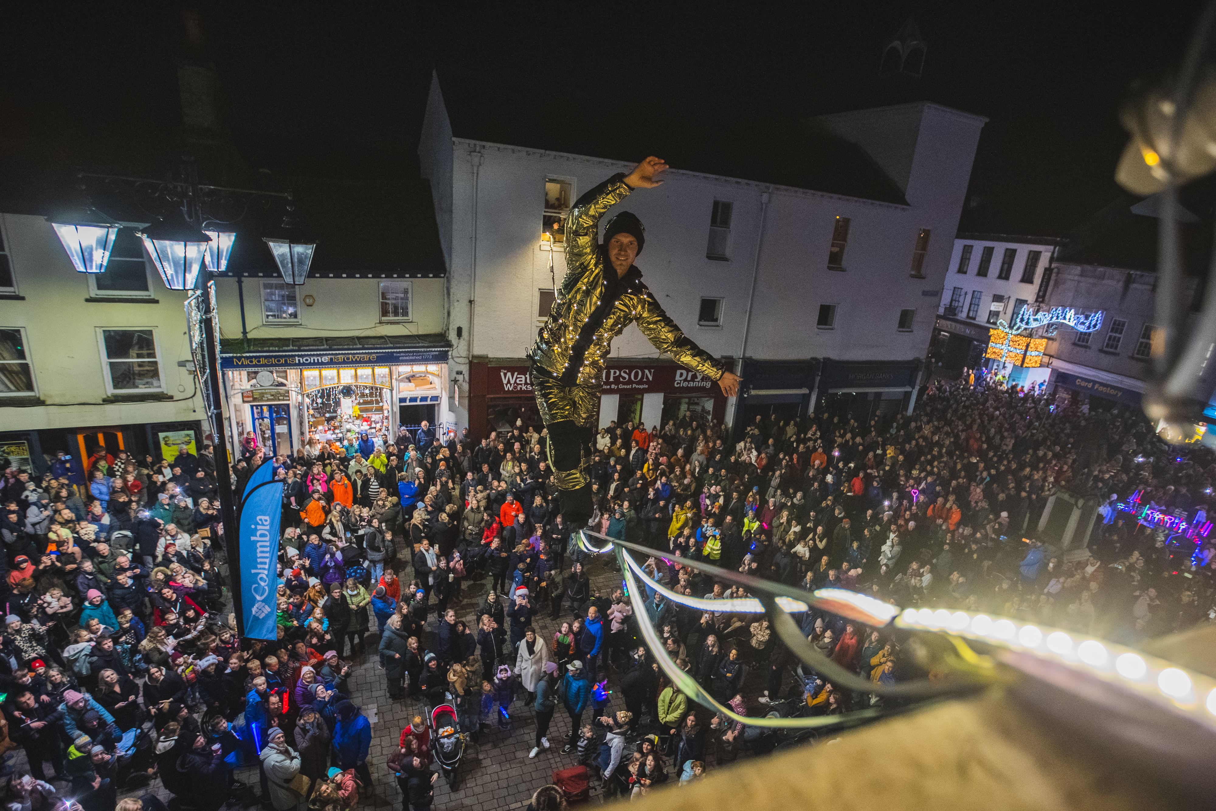 Large crowd gathered in town square at night, surrounded by white buildings with shops. Colourful clothing, bright street lighting.