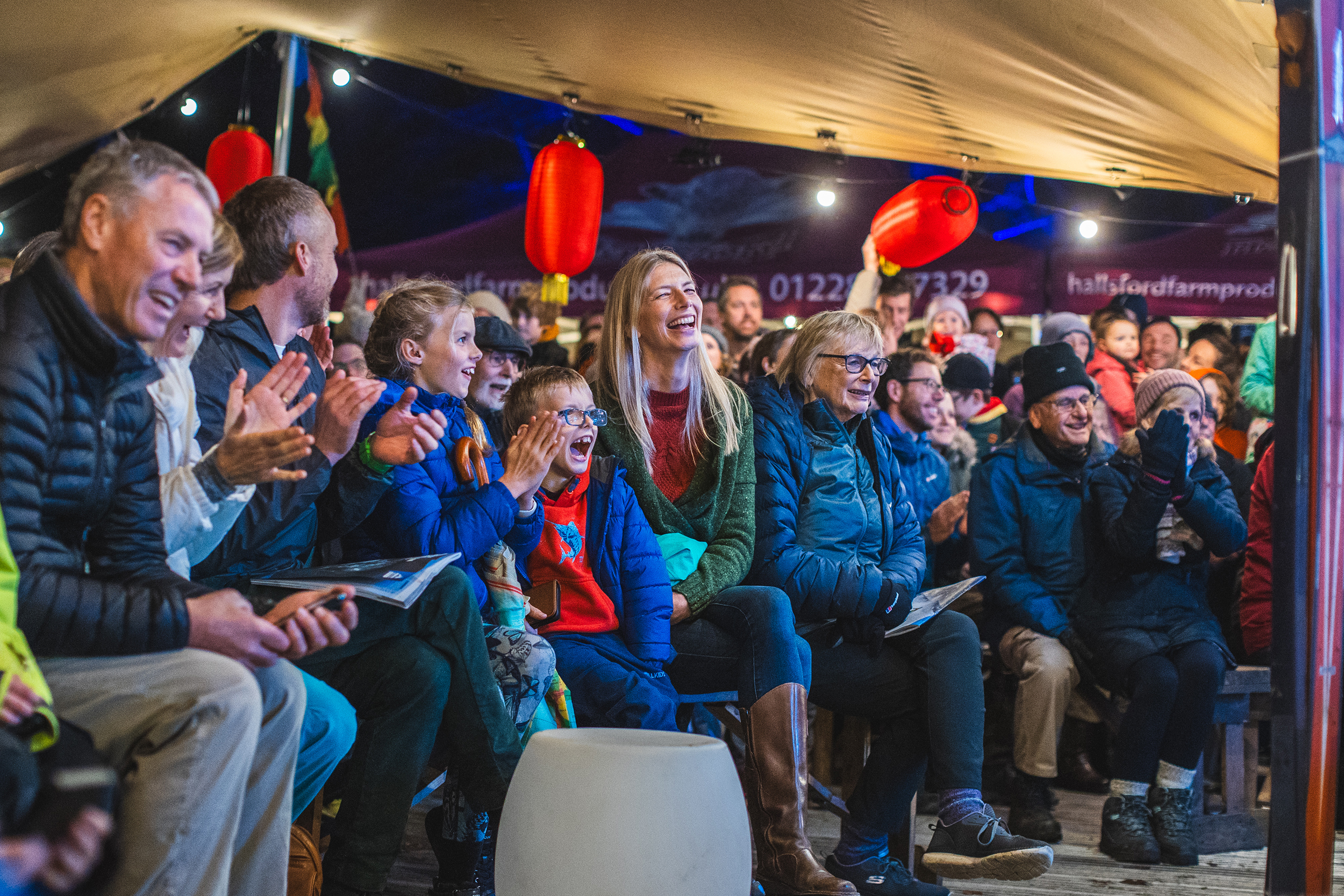 Crowd of people laughing and clapping whilst seated under tent with red lanterns and warm lighting overhead.