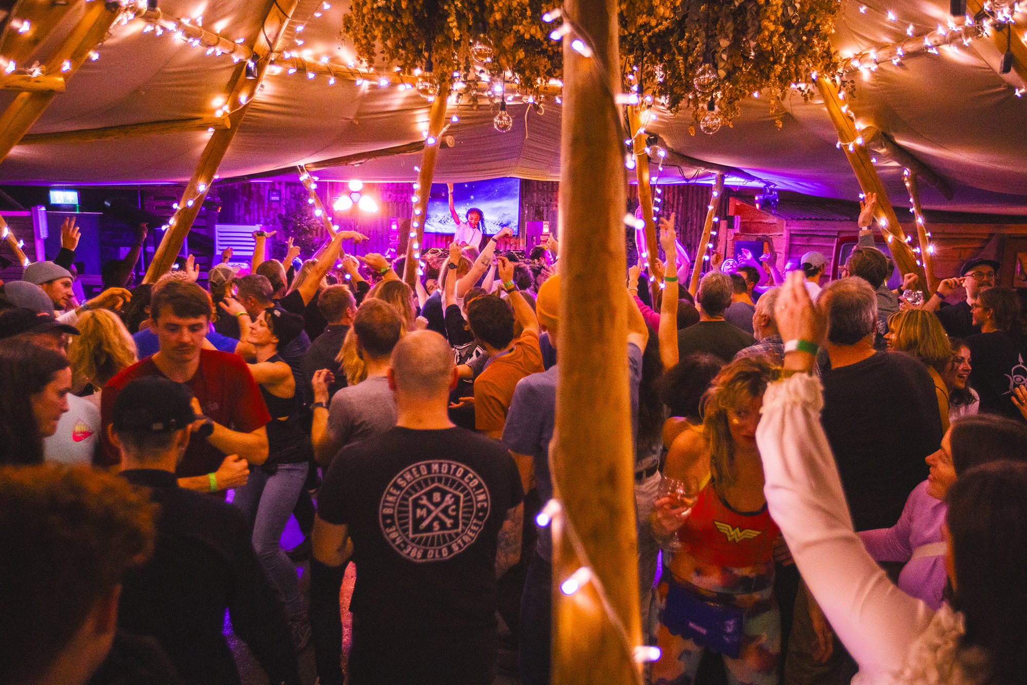 Crowded indoor party venue with people dancing under warm string lights and purple stage lighting, wooden posts visible.