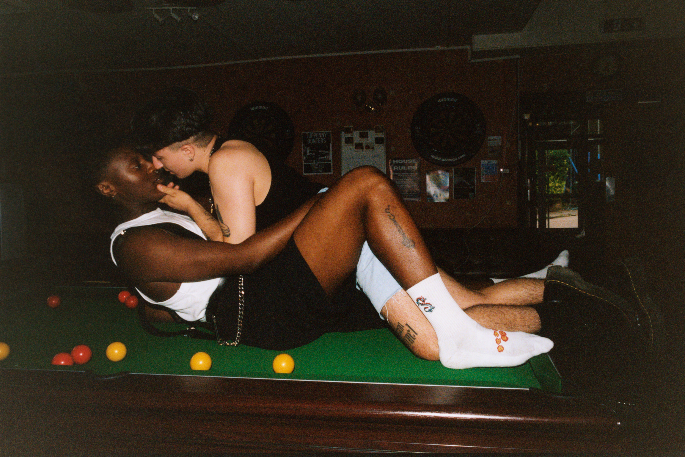 Woman in white bikini lying on green snooker table with coloured balls scattered around in dimly lit pub interior.