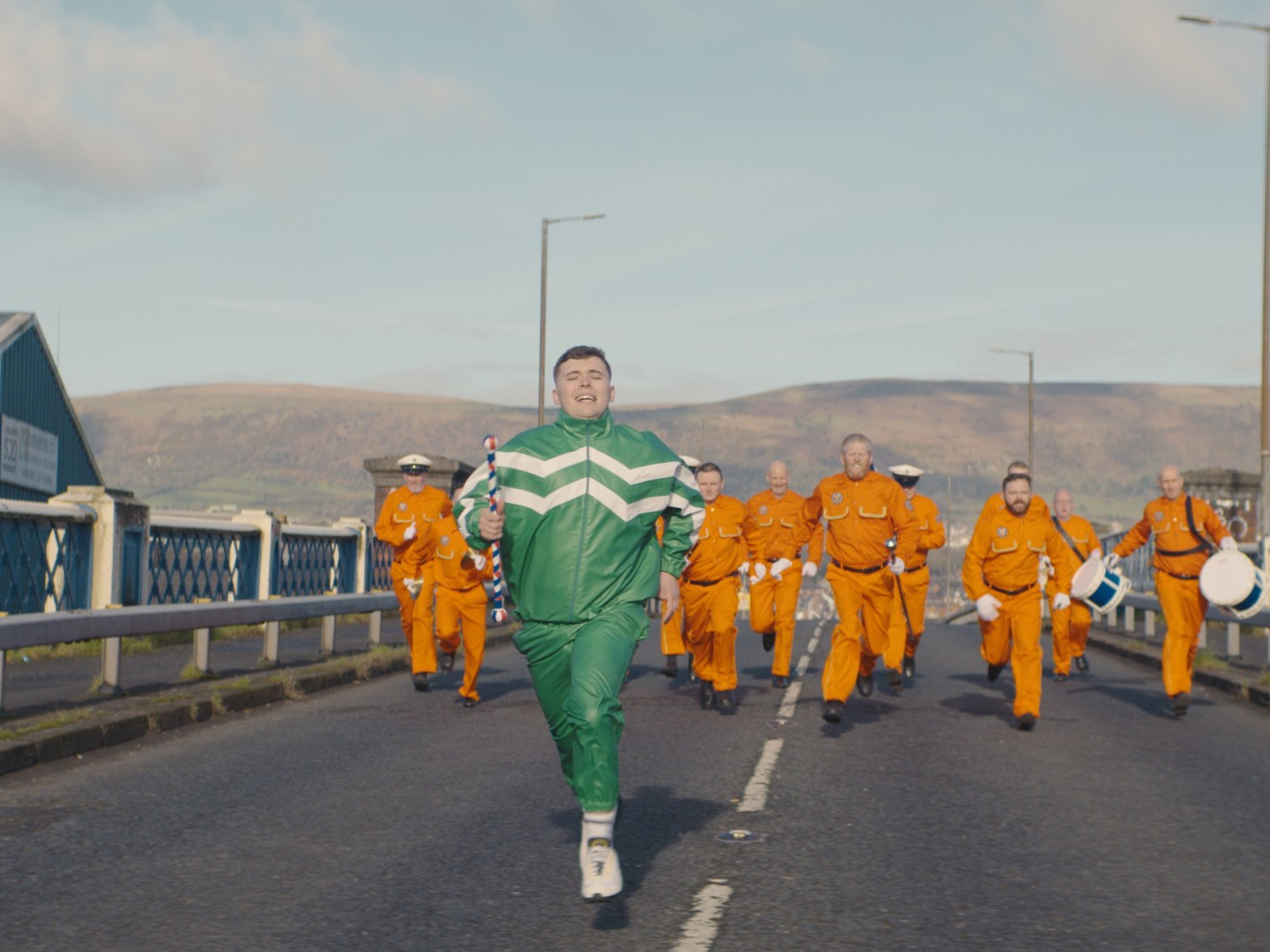 A group of people wearing orange and green uniforms walking on a road with hills in the background.