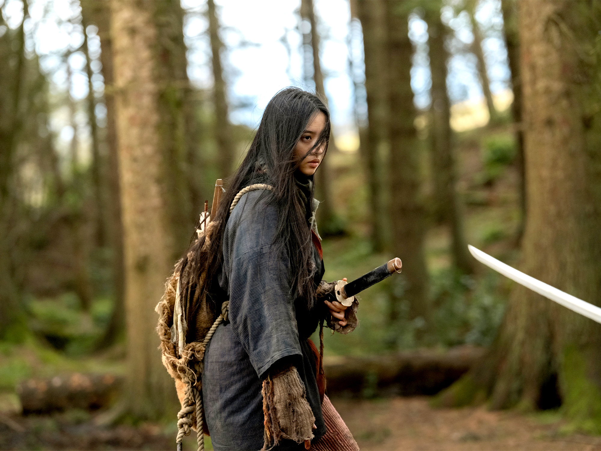 A person in a dark grey cloak holding a sword stands in a forested area with tall trees.