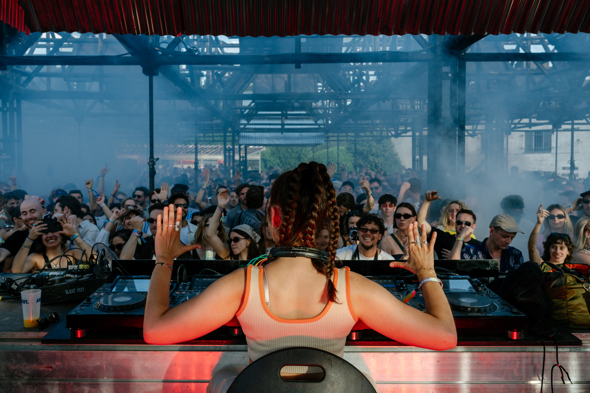 Crowded electronic music event with a female DJ on stage, raising her hands and facing away from the camera, surrounded by an enthusiastic crowd.