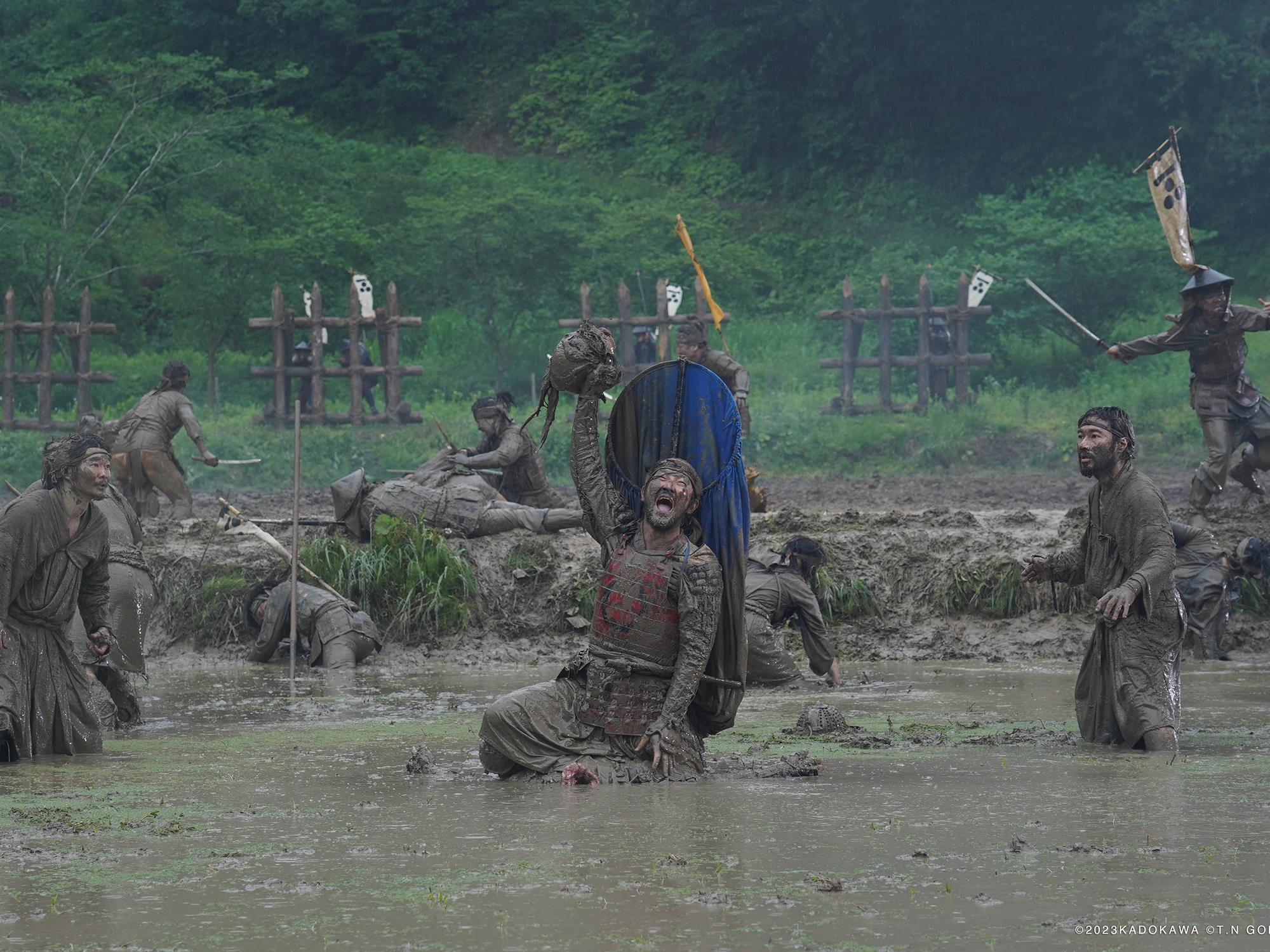 Samurai in traditional uniform, covered in mud, standing in a muddy river with trees and foliage in the background. One of them holds a human head aloft.