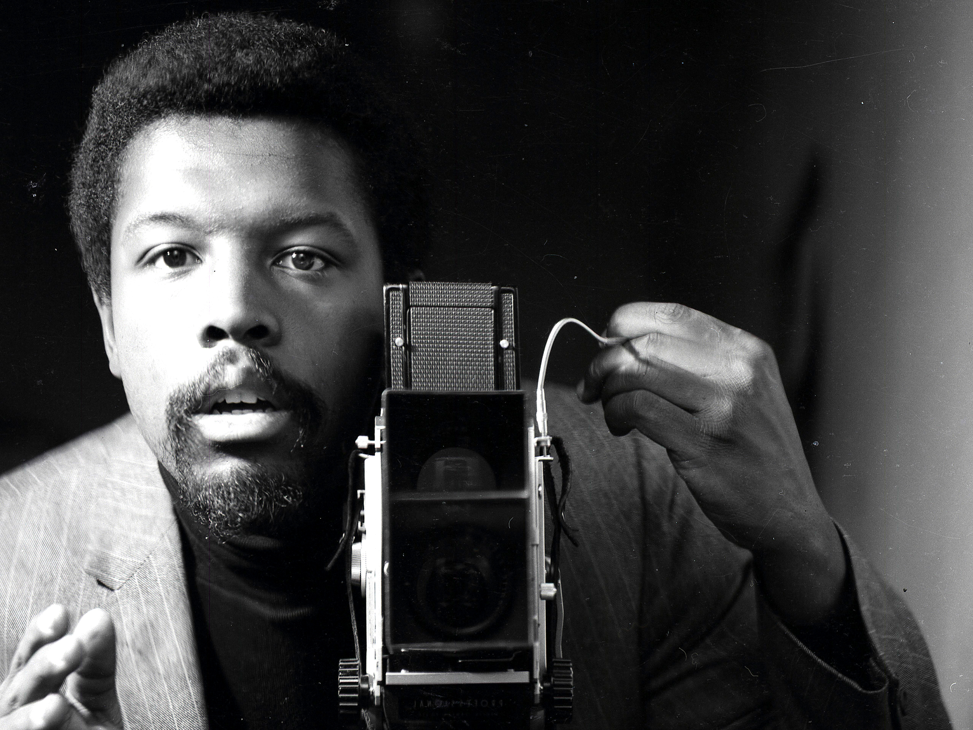 Black and white portrait of a bearded Black man holding a vintage folding camera, positioned close to his face in dramatic lighting.